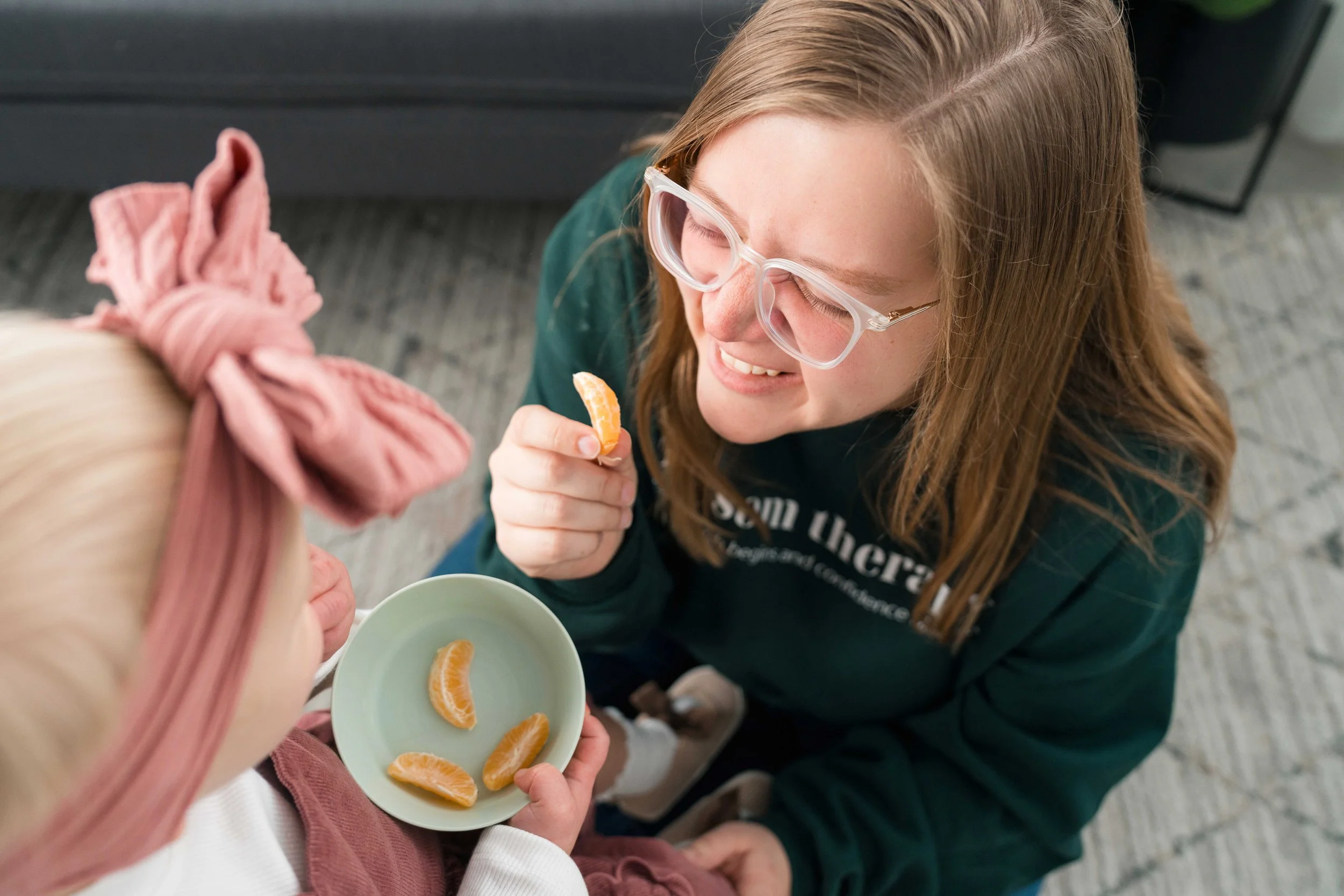A woman with glasses smiling while offering a peeled orange piece to a child with a pink headband. The child is holding a small plate with four orange slices.