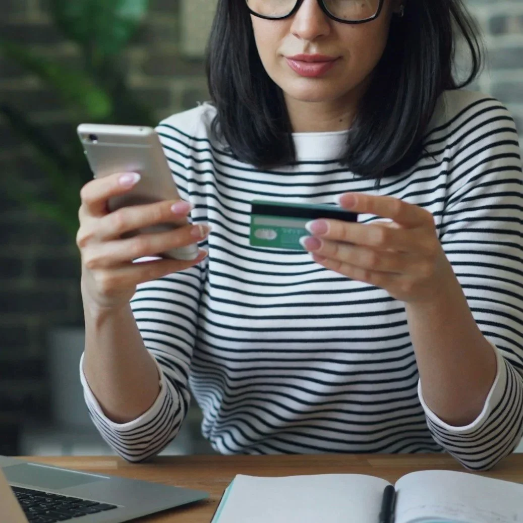 A woman with black hair and glasses wearing a striped shirt, holding a smartphone in one hand and a credit card in the other, sitting at a desk with a laptop, notebook, and pen.
