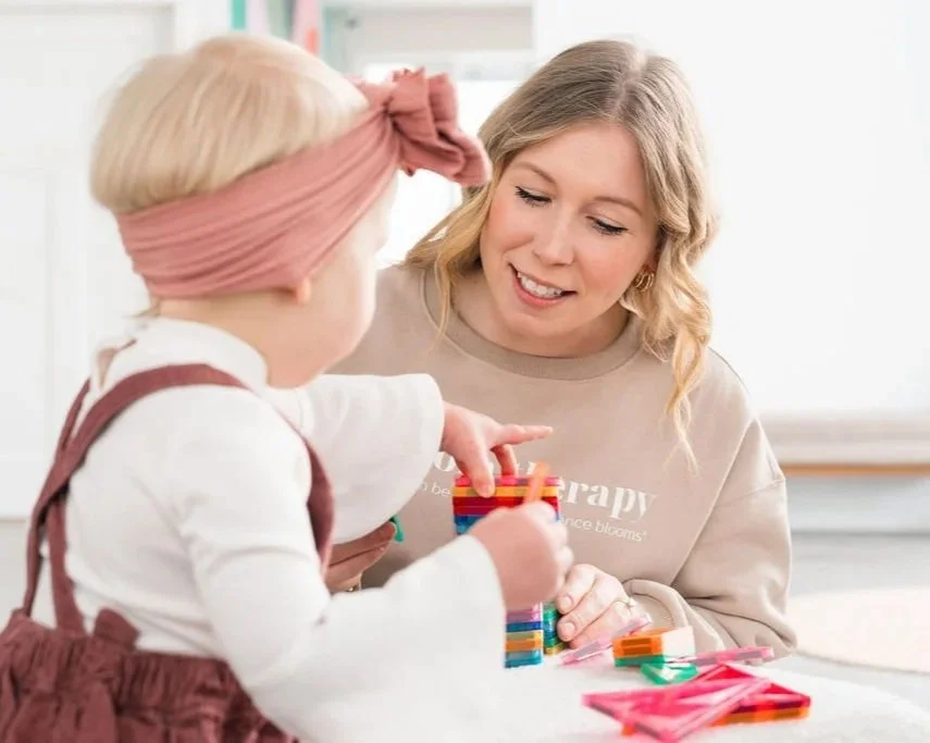A woman smiling while playing with a young girl who has a pink headband, in a bright room, with building blocks on the table.