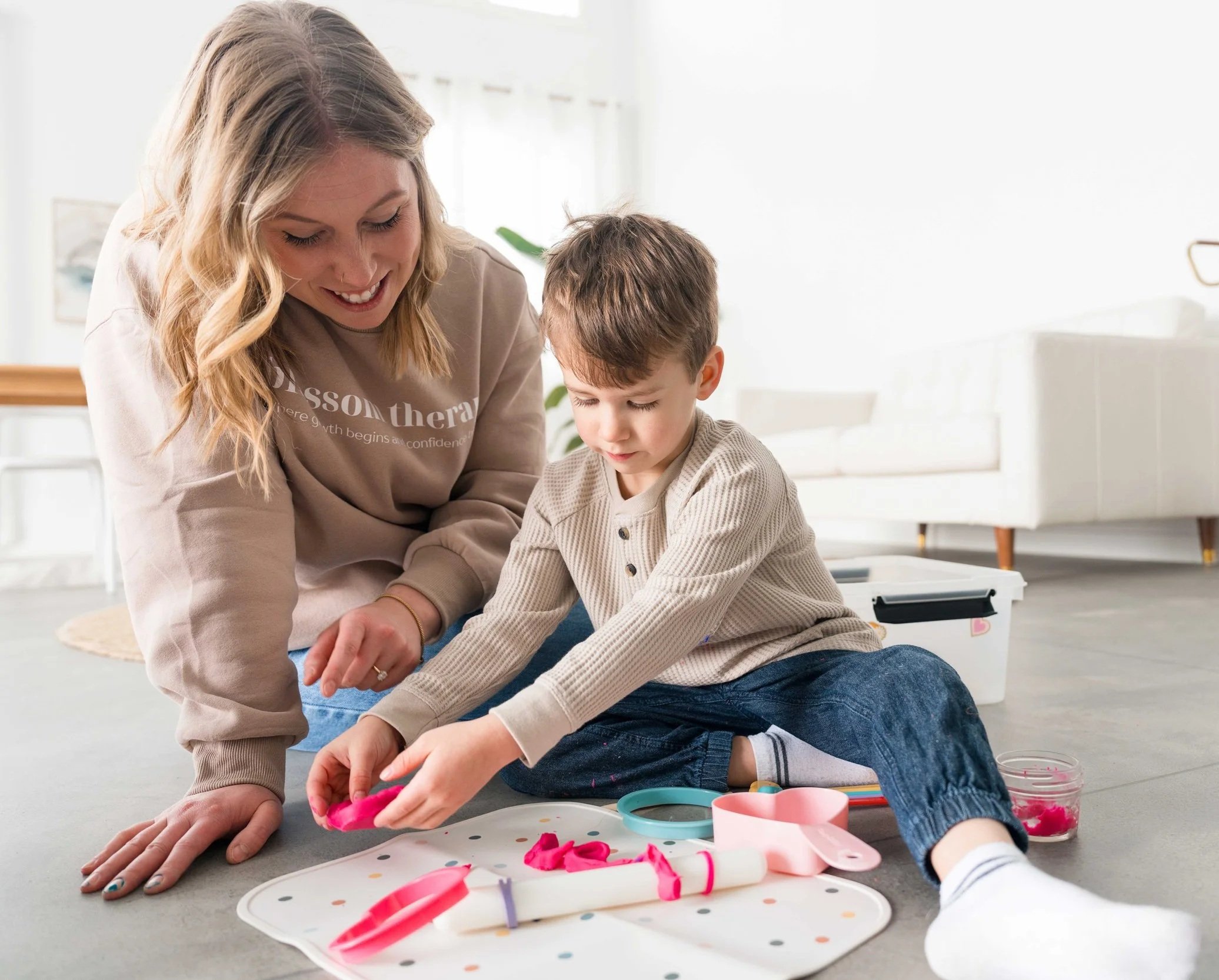 A woman and a young boy sitting on the floor playing with pink modeling clay, surrounded by baking tools and a white tray with colorful dots, in a bright living room.