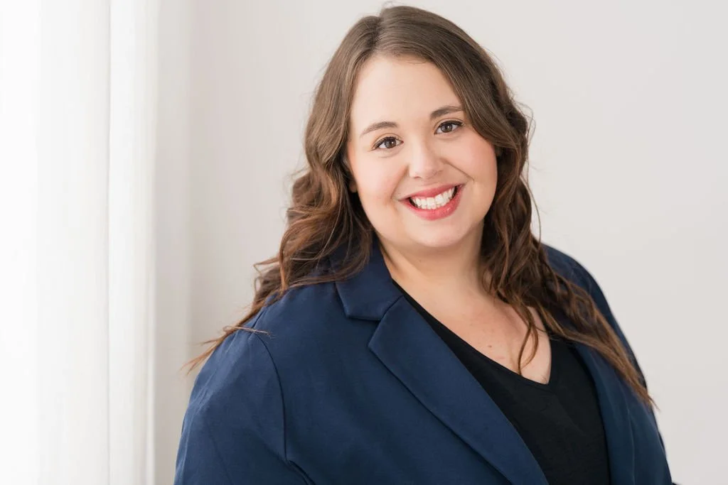 A woman with long brown hair, smiling, wearing a dark blazer over a black top, standing against a light-colored wall.