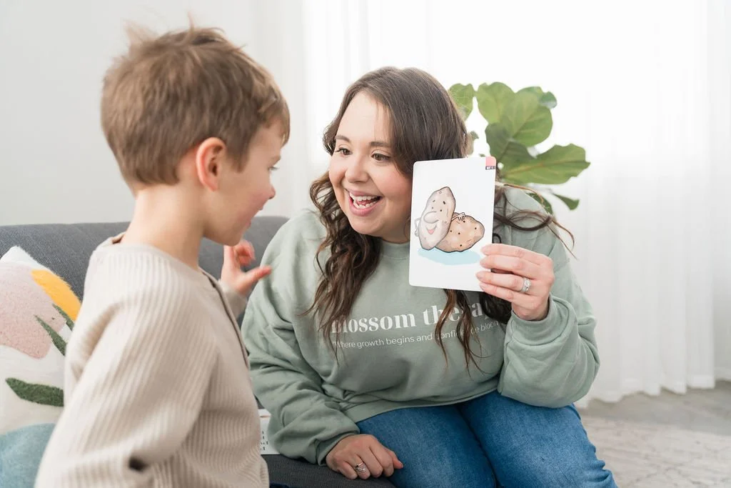 A woman smiling and holding a card with a drawing of potatoes, talking to a young boy sitting on a couch.