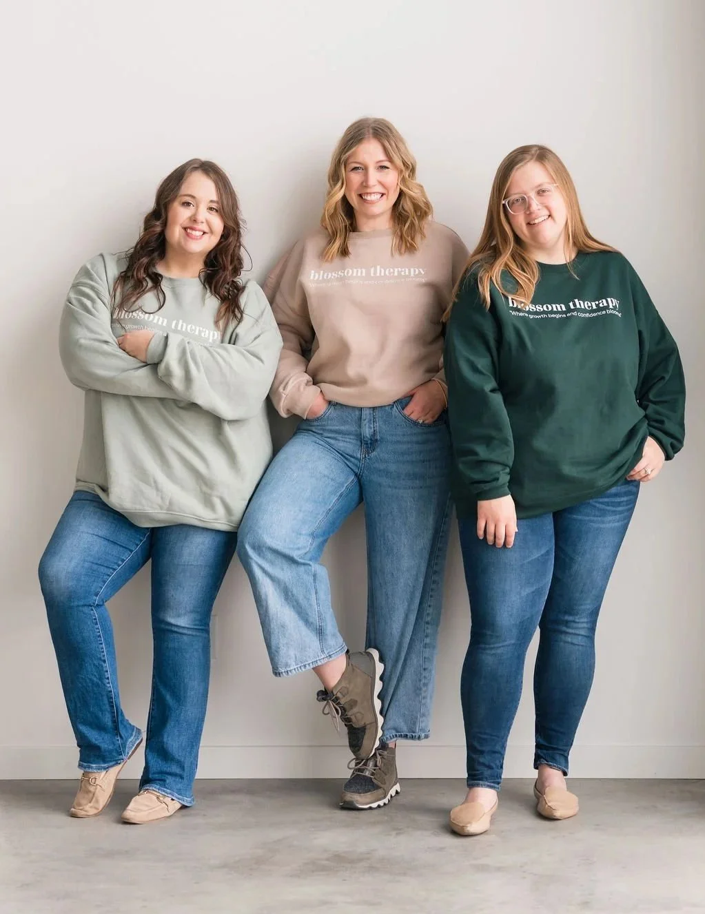 Three women smiling and posing against a plain white wall, wearing sweatshirts with 'blossom therapy' written on them.
