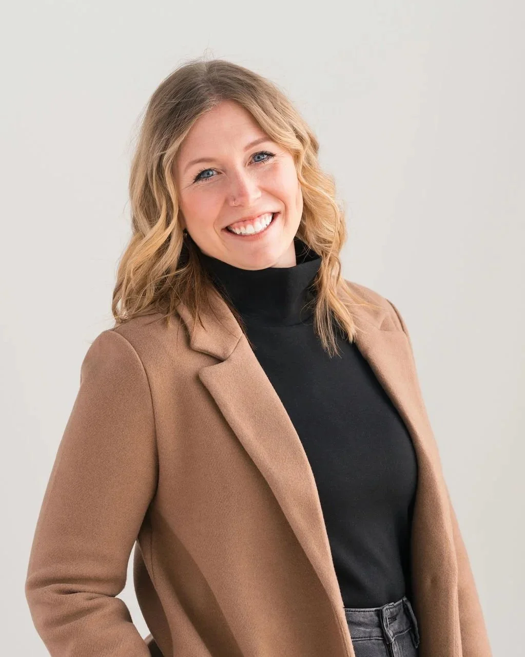 A smiling woman with wavy blonde hair wearing a black turtleneck and a tan blazer, standing against a plain white background.