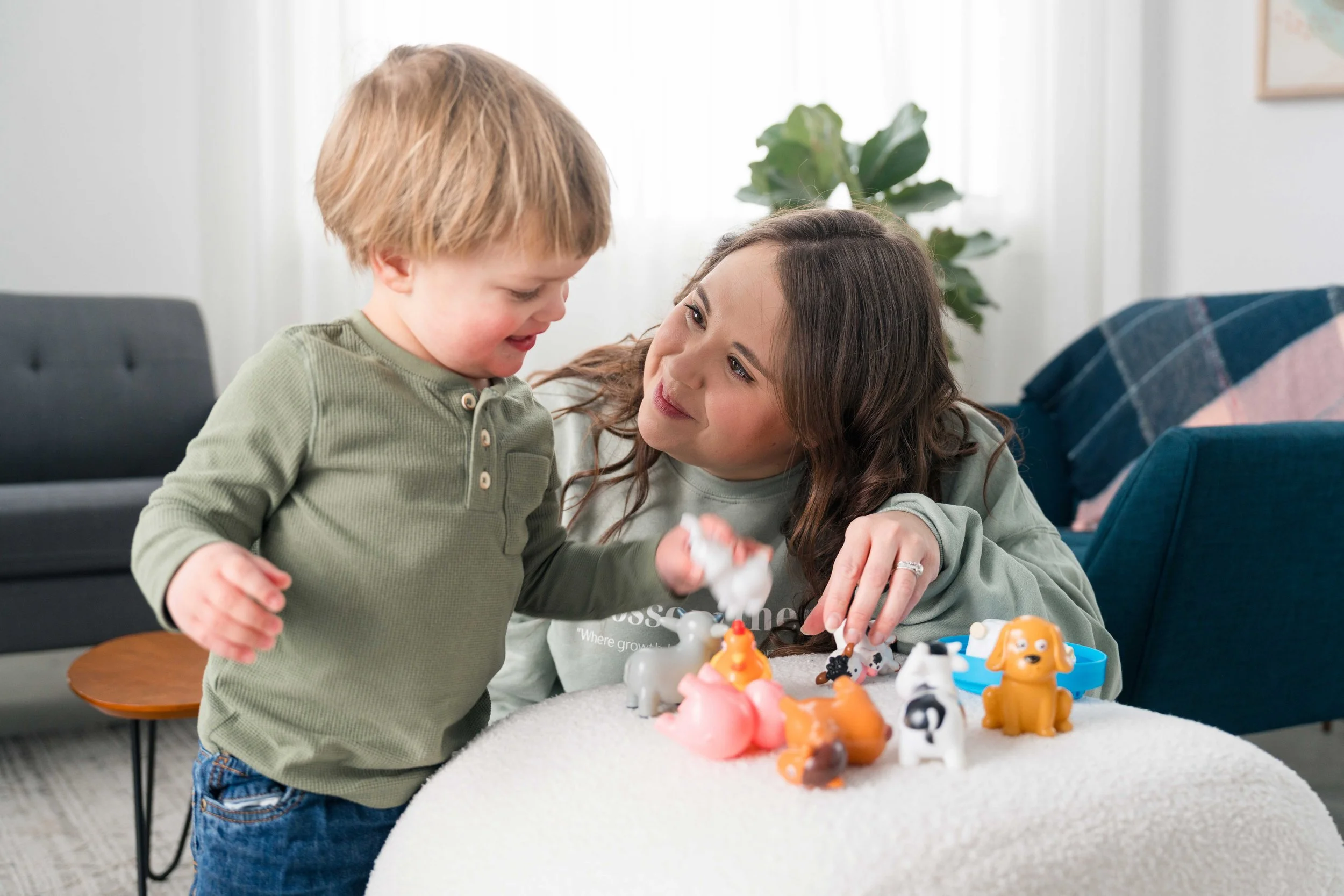 A woman and a young boy playing with toy animals at a table in a bright living room.