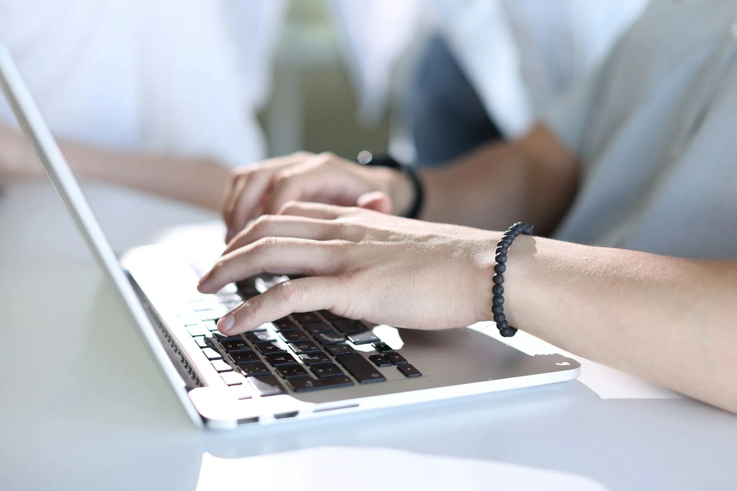 Close-up of a person's hands typing on a silver laptop keyboard with a black beaded bracelet on the wrist, sitting at a white desk.