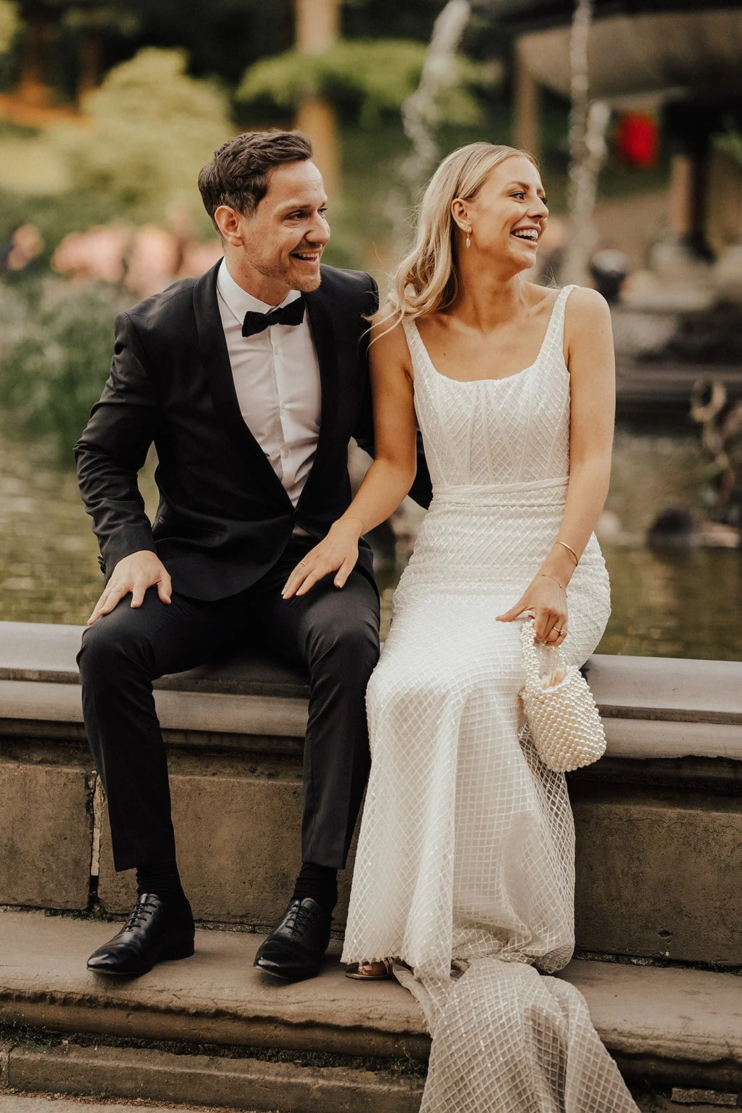A bride and groom sitting on a ledge near a fountain, smiling and laughing. The groom is wearing a black tuxedo with a bow tie, and the bride is dressed in a white, textured wedding gown, holding a pearl-like clutch, with blurred greenery in the background.
