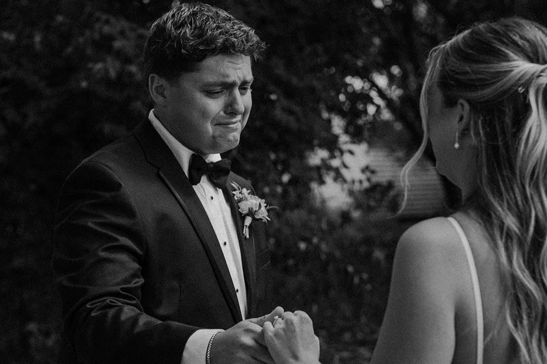 A groom in a tuxedo holding a bride's hand, both are emotional during their wedding ceremony outdoors.