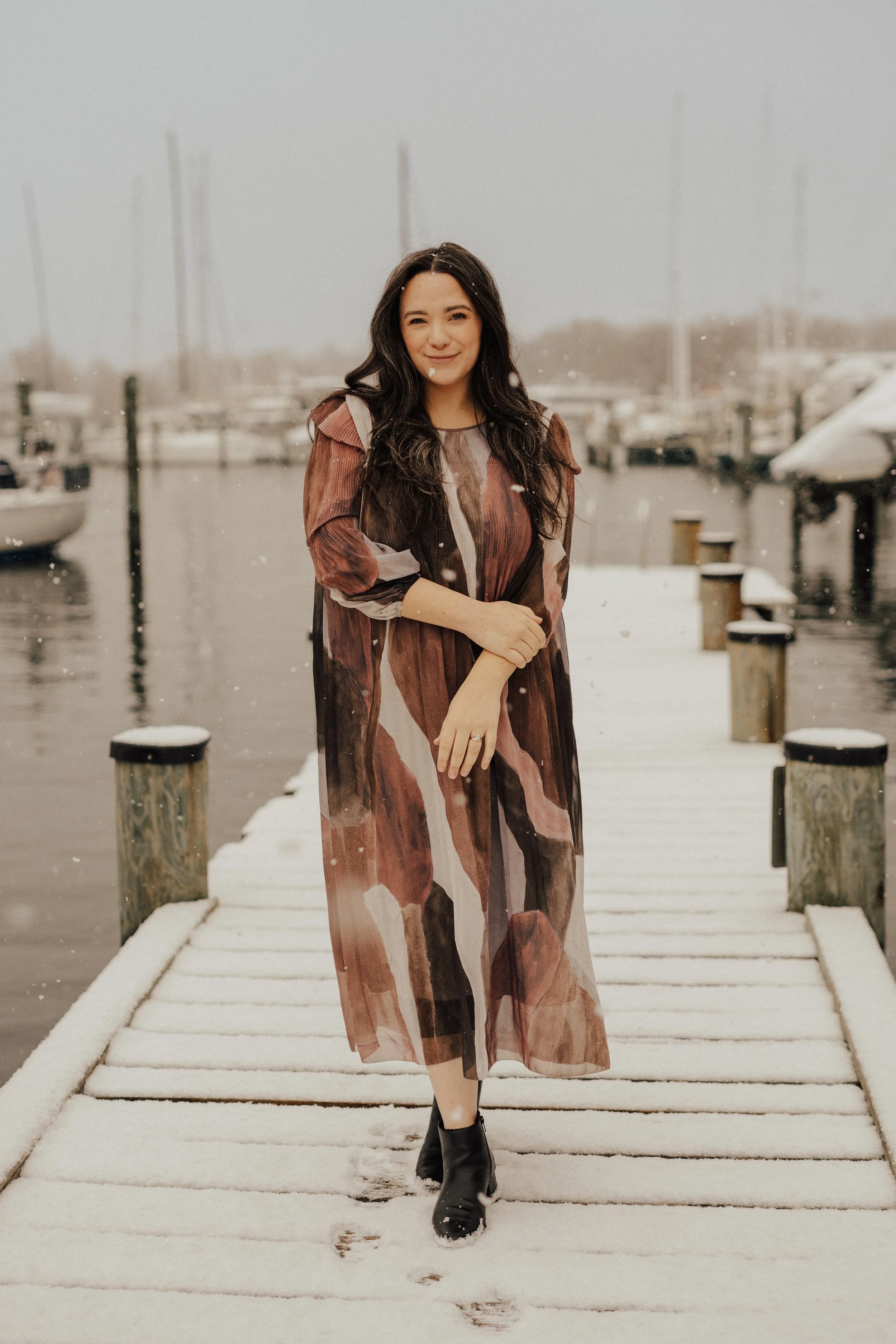 A woman in a long, patterned dress and black boots standing on a snow-covered dock by the water, with boats and a cloudy sky in the background.