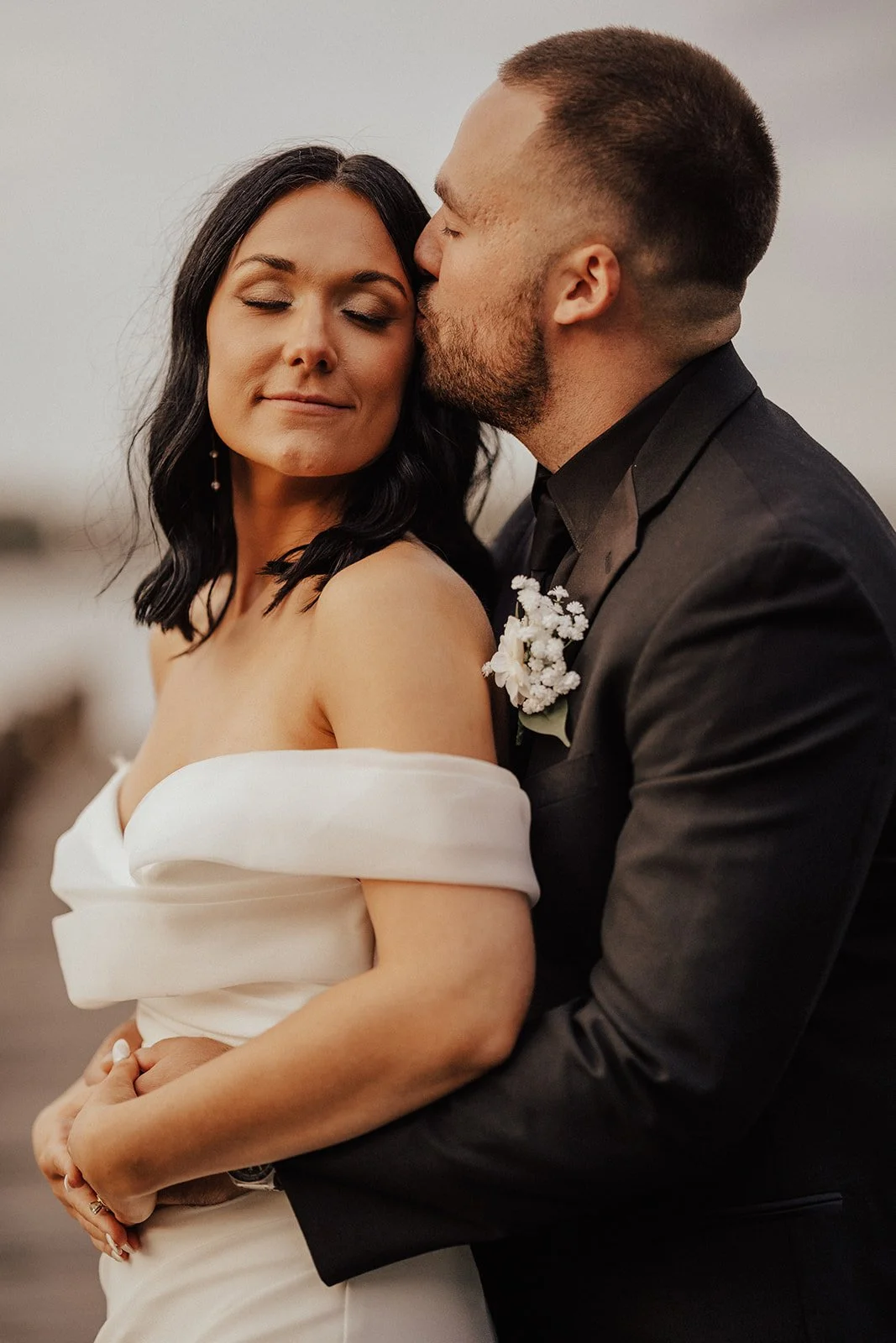 A couple in formal attire, with the man kissing the woman on her forehead. The woman has dark, wavy hair and is wearing an off-shoulder white dress. The man has short hair and a beard, and is dressed in a black suit with a boutonniere of white flowers. The background is an outdoor setting.