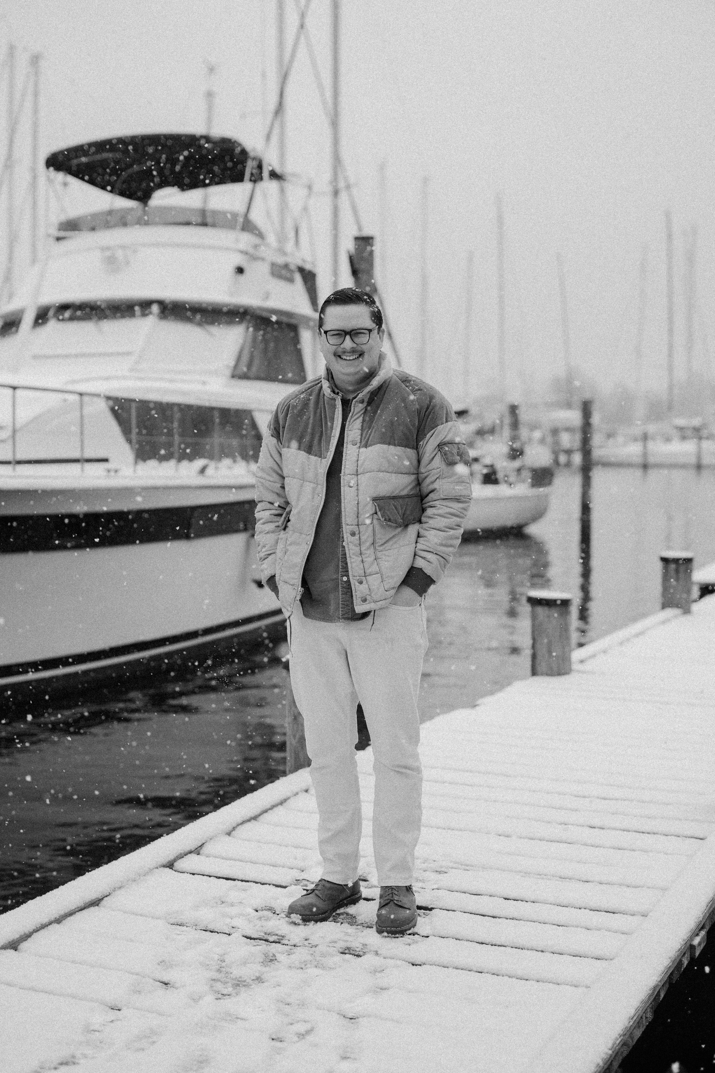 A man standing on a snow-covered dock with his hands in his pockets, smiling, with boats docked behind him amid falling snow.