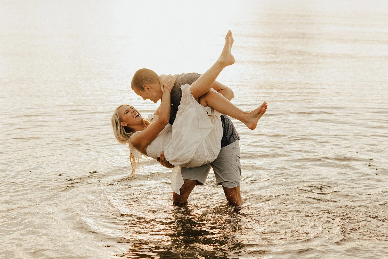 A happy woman and man are in shallow water at the beach, with the man lifting the woman in an embrace. The woman is wearing a white dress, and both are smiling and laughing.