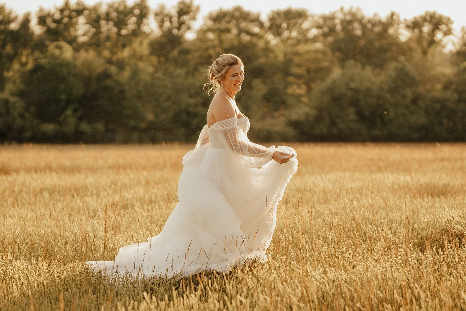 A woman in a white dress standing in a wheat field during sunset, smiling and holding her dress.