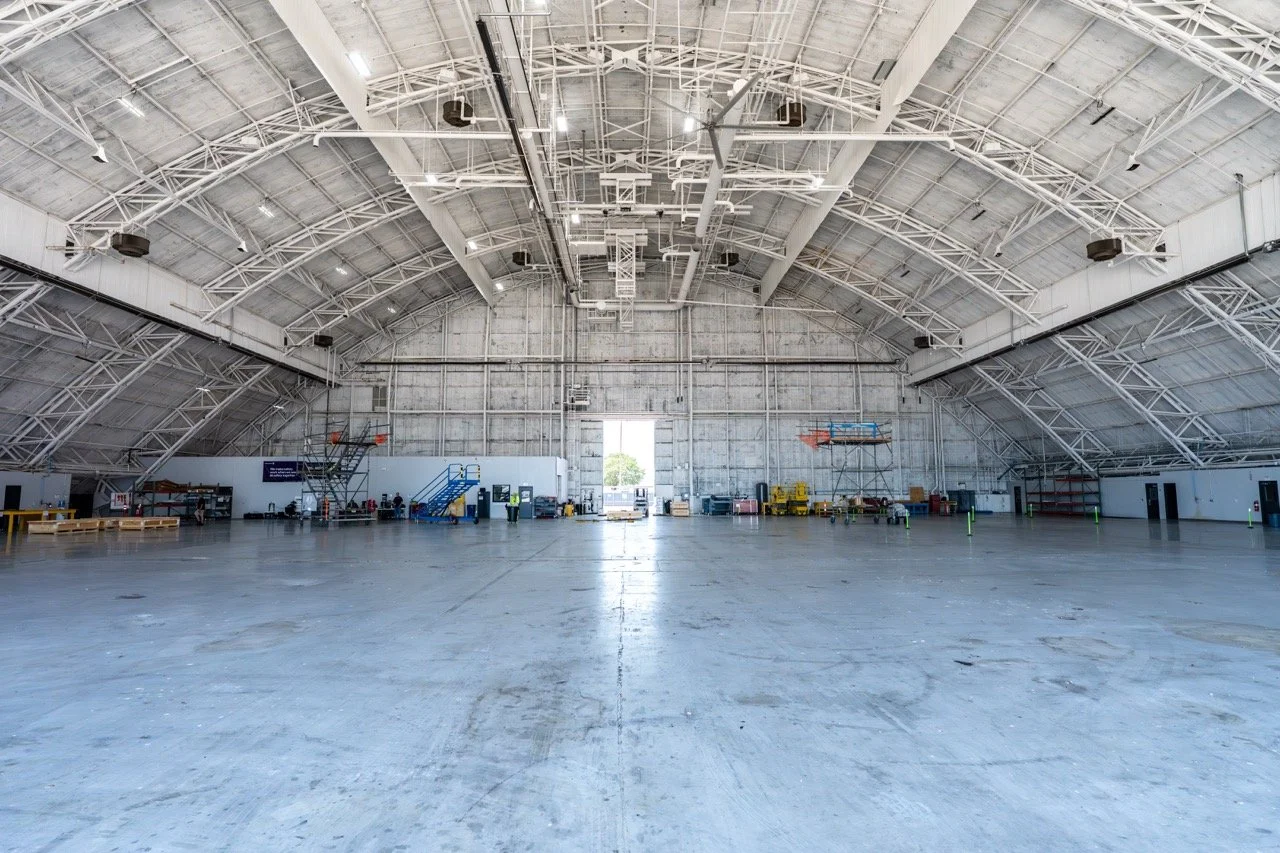 Interior of an empty hangar with a high arched ceiling, exposed metal beams, and large open entrance at the back, with some equipment and personnel along the back wall.