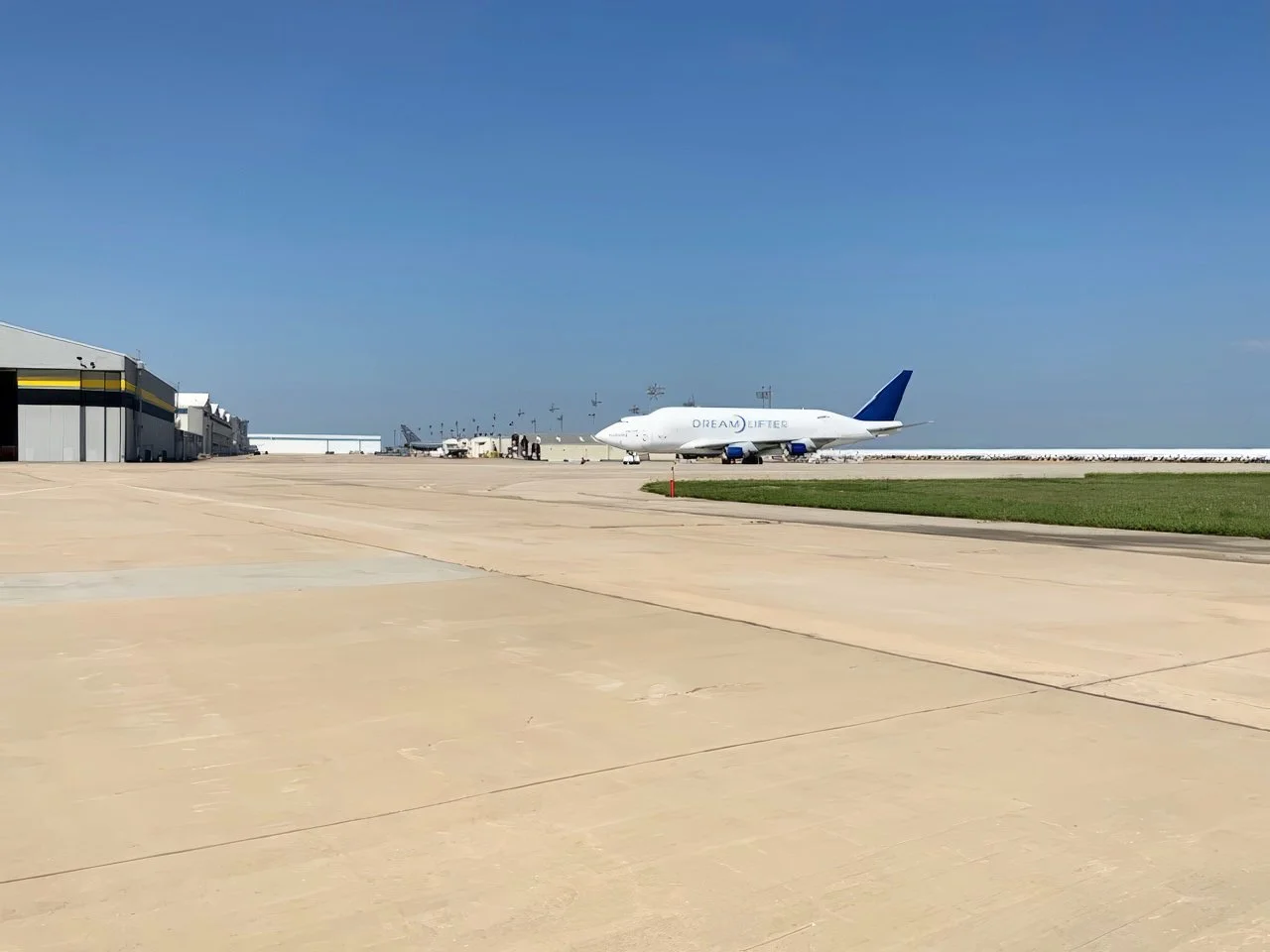 A large airplane with the words 'Dream Lifter' on the side is parked on the tarmac near airport hangars, under a clear blue sky.