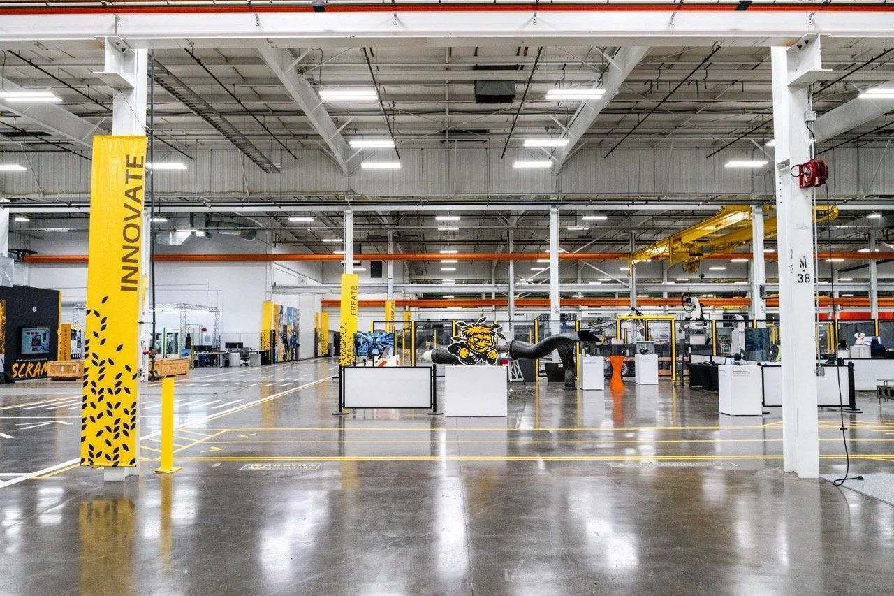 Empty industrial manufacturing or warehouse space with bright overhead lighting, yellow and black safety and navigation signs, and various machinery and workstations in the background.