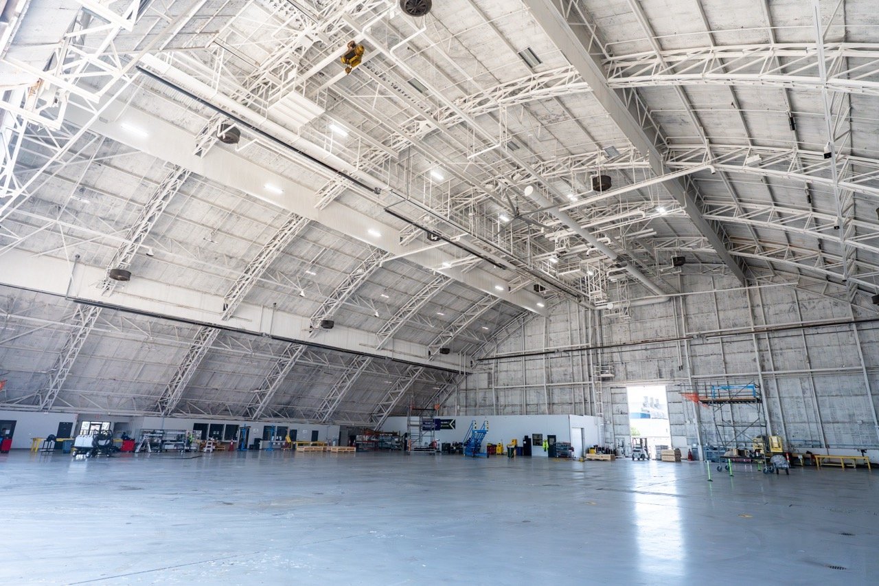 Empty aircraft hangar with high arched ceiling and industrial lighting, tools, and equipment along the walls.