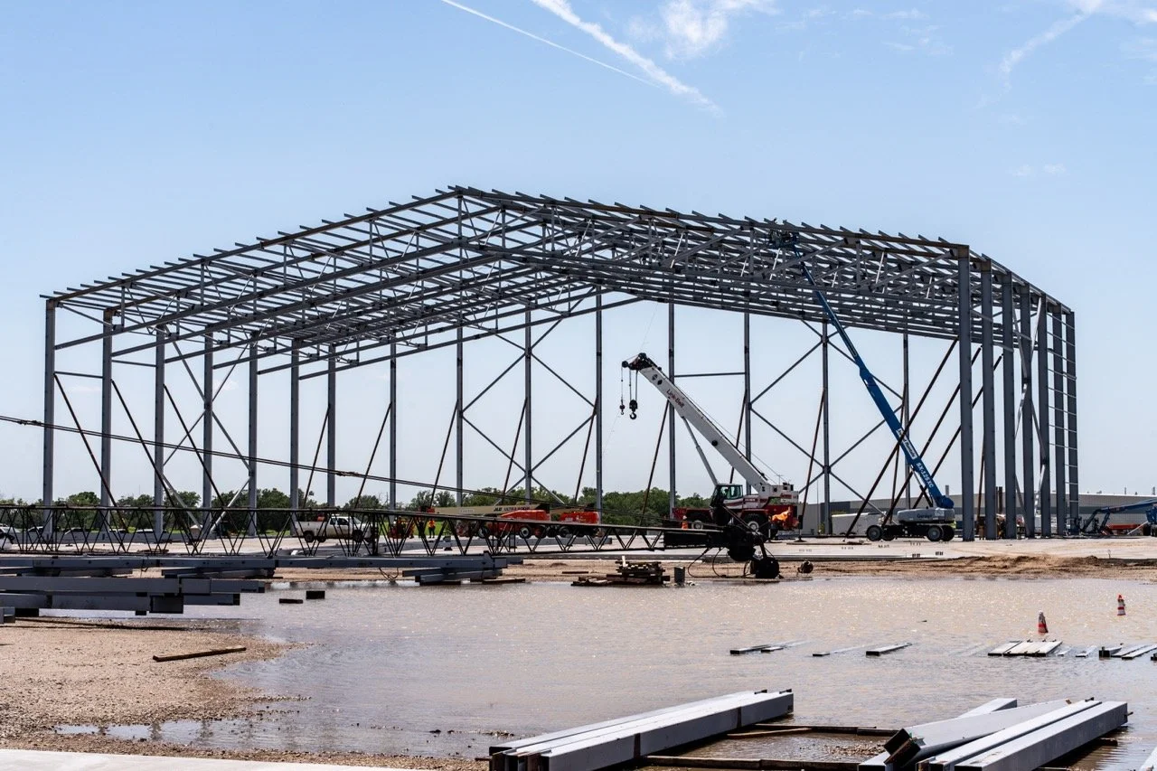 Steel framework of an airport hangar under construction with construction equipment and materials on the ground, clear sky in the background.