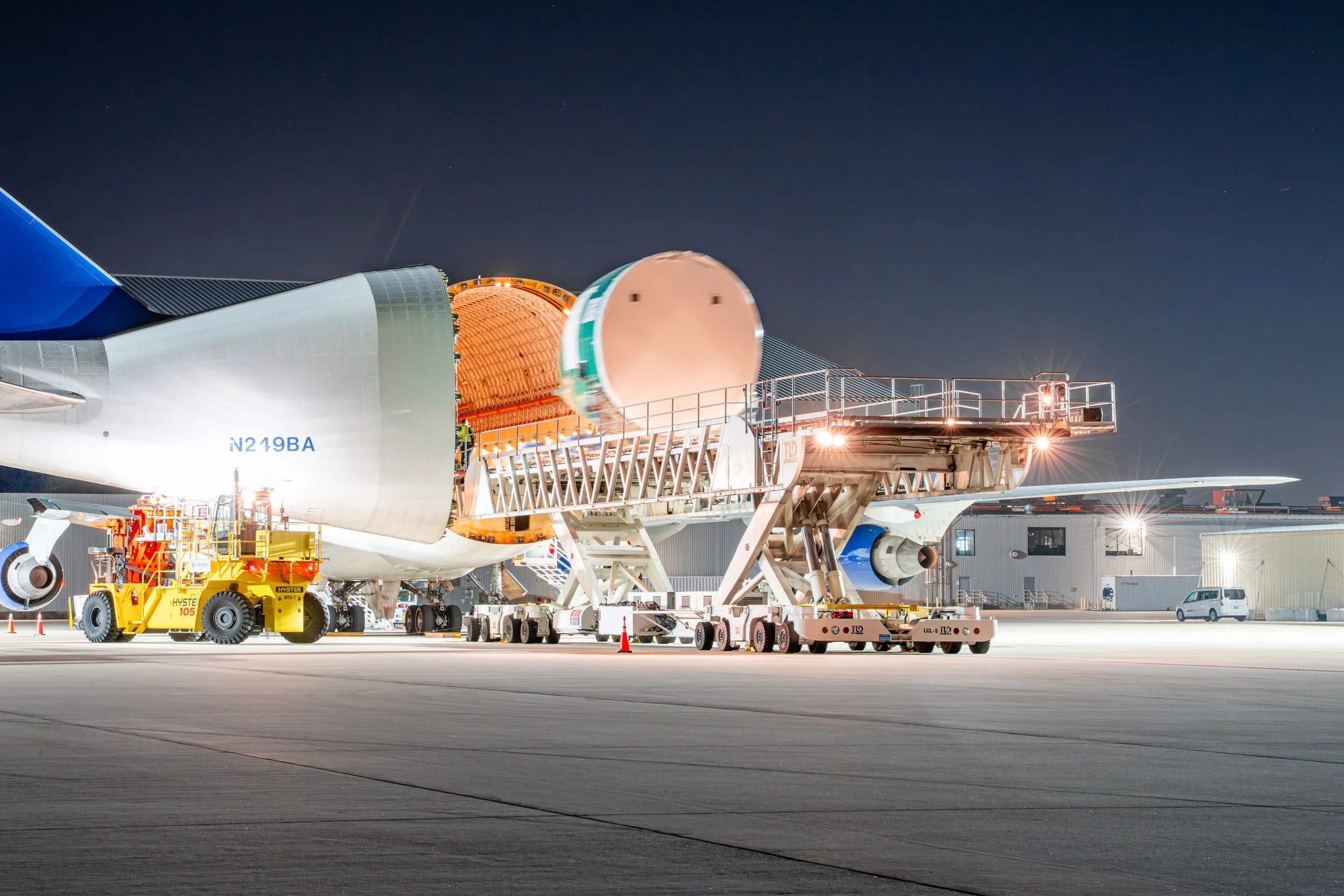 A large commercial airplane at night being loaded or serviced with ground support equipment, with its engine cowling open and ramp connecting to the aircraft.
