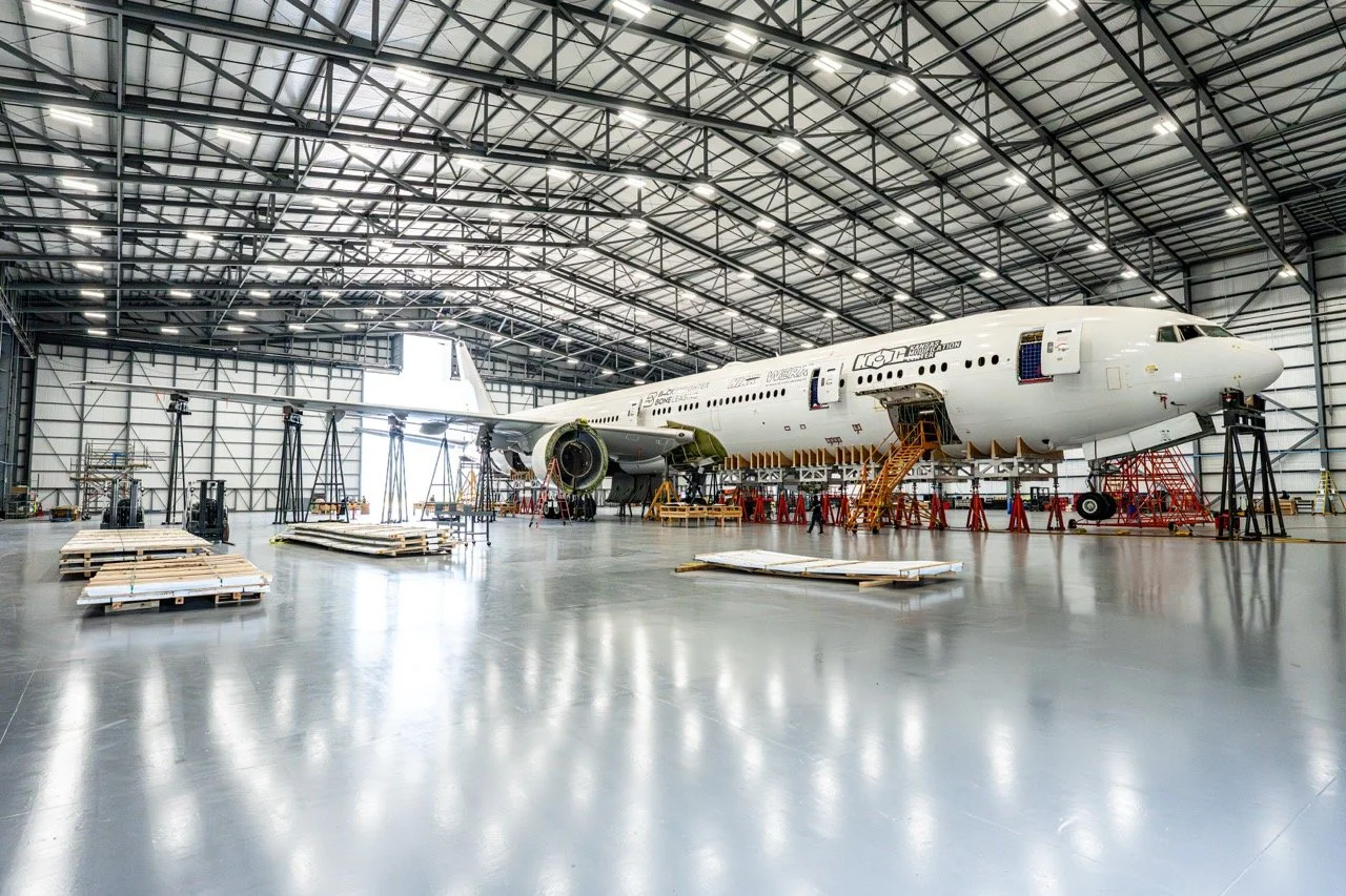 A large airplane under maintenance inside a spacious aircraft hangar with a high ceiling and bright lighting, surrounded by equipment and walkways.