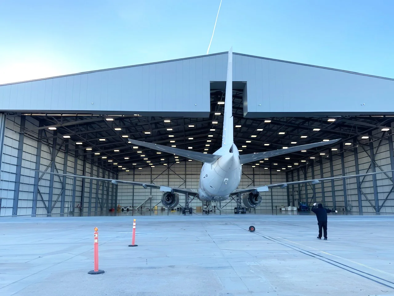 A large white airplane inside an aircraft hangar with open doors. There is a person on the right side taking a photo and several cones on the ground.