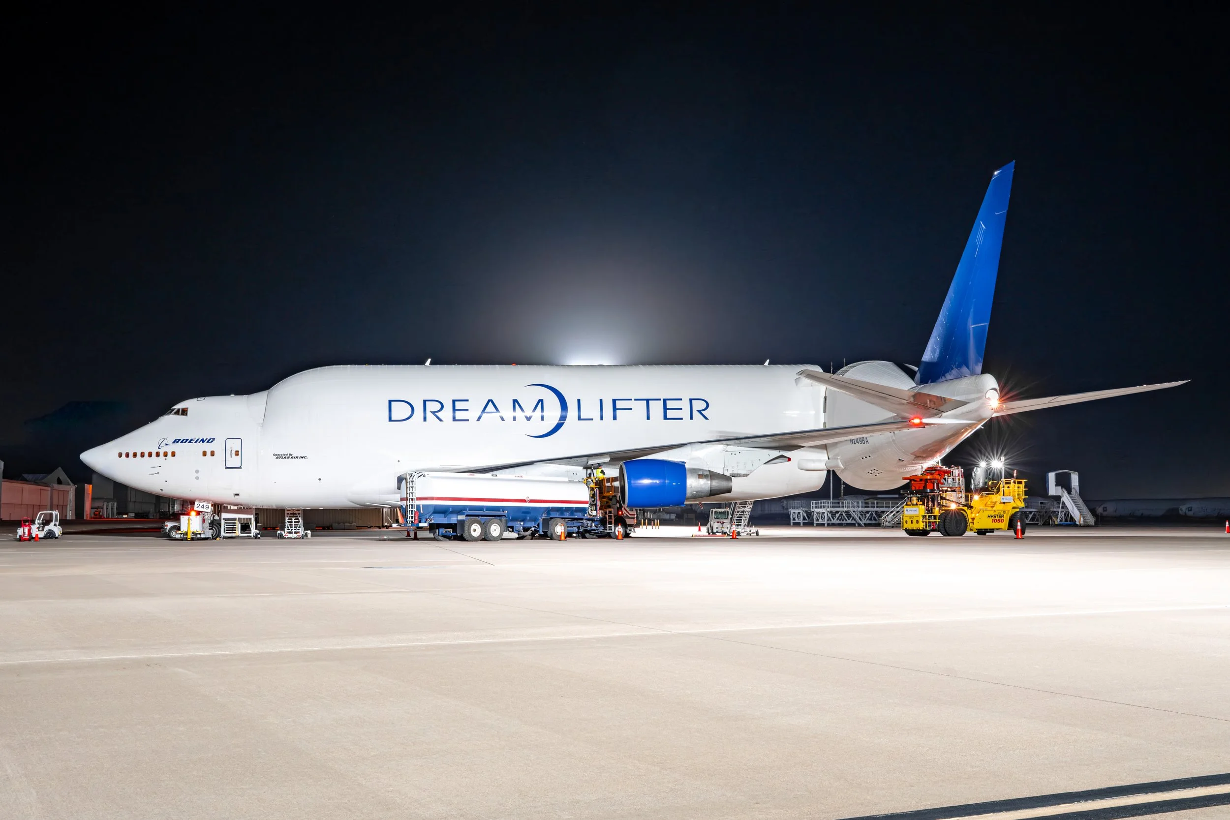 Nighttime view of a large Boeing Dreamlifter cargo aircraft on the tarmac, connected to fueling trucks and ground support equipment, with a dark sky in the background.