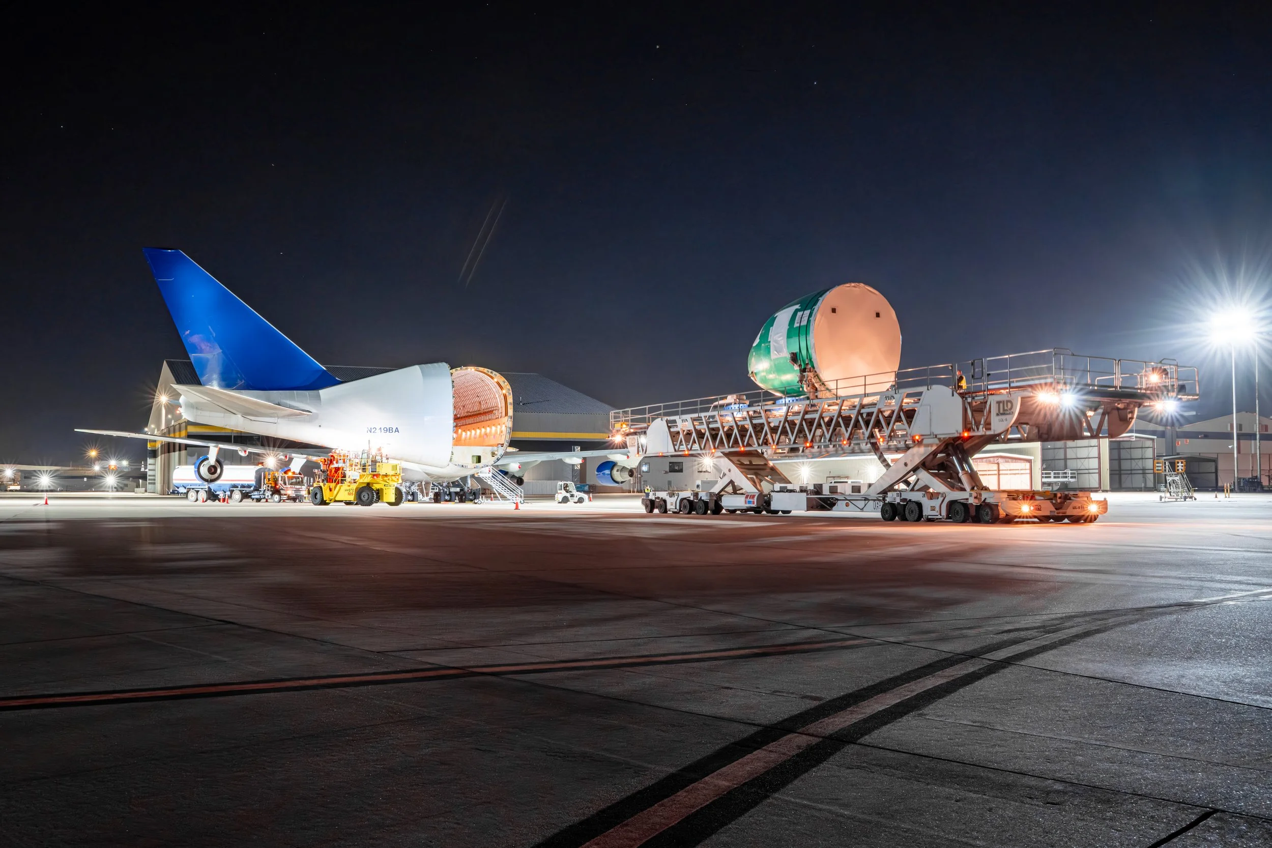 A Dreamlifter aircraft on the tarmac at night with a cargo loader unloading a large, cylindrical green and white object from the plane.