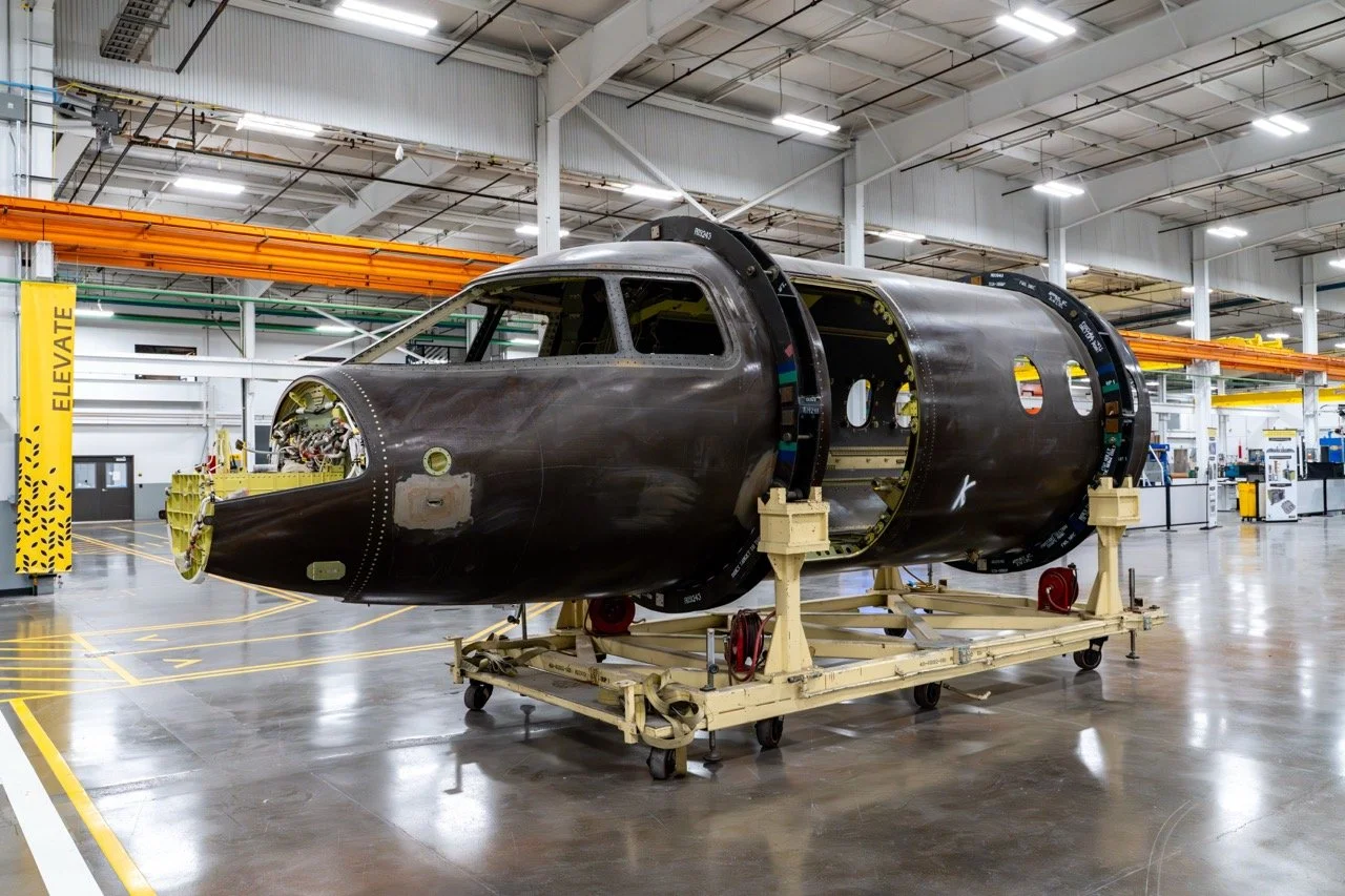 Aircraft fuselage in an assembly facility, placed on a movable support frame, with the nose and cockpit area visible but lacking windows and engines.