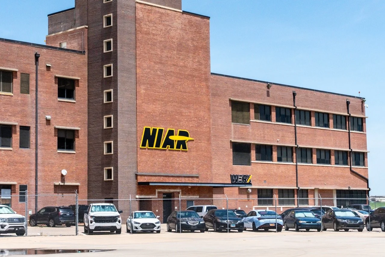 A large brick building with the NIAR logo on the side. There are parked cars in front of the building and a chain-link fence separates the parking lot from the building. The sky is clear and blue.