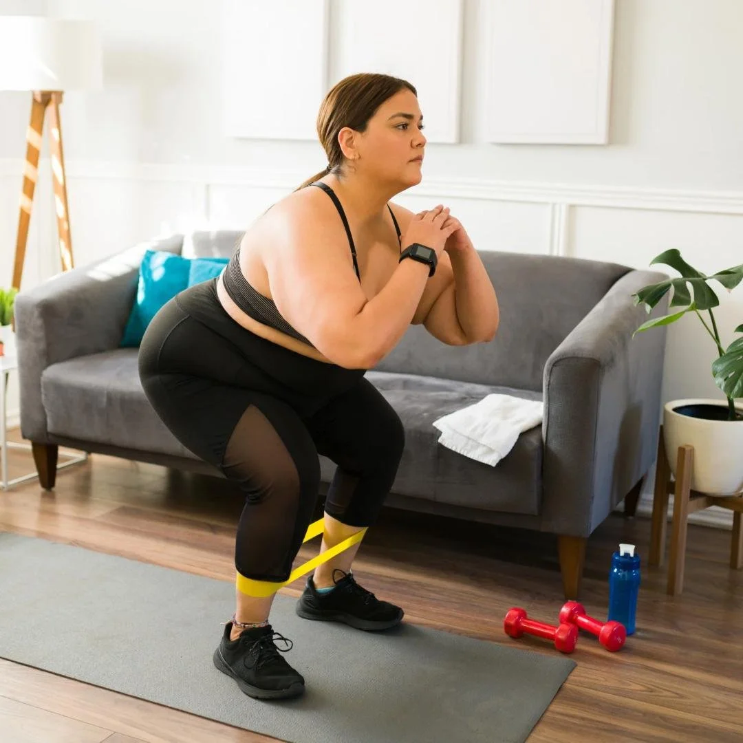 Mujer haciendo ejercicio en casa con banda de resistencia, en posición de sentadilla, ropa deportiva, en un espacio interior con sofá y plantas.