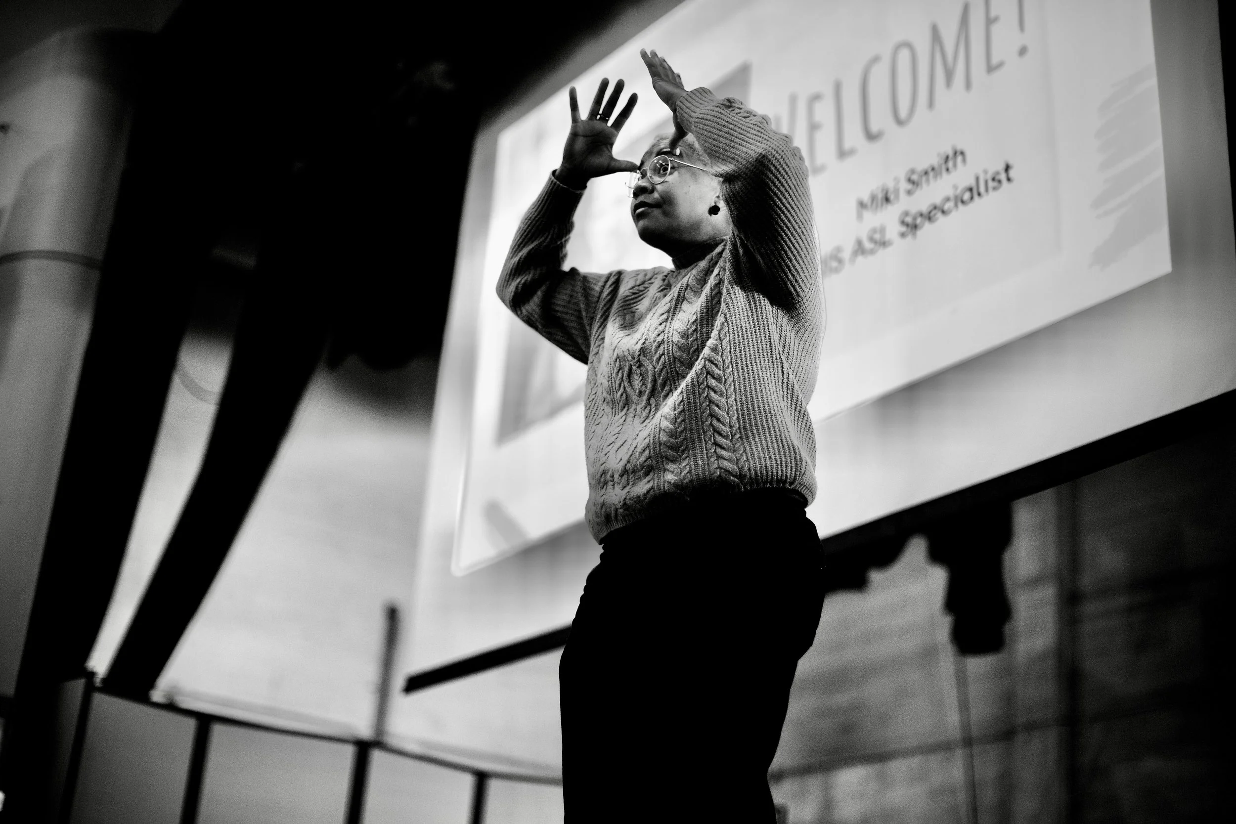 Black-and-white photo of Miki standing in front of a projected screen, signing with both hands raised near their face during a presentation.