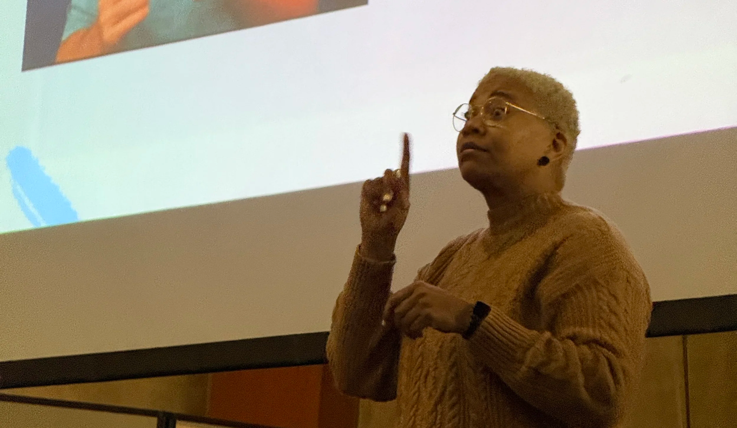 Miki stands at the front of a room, signing while facing the audience.