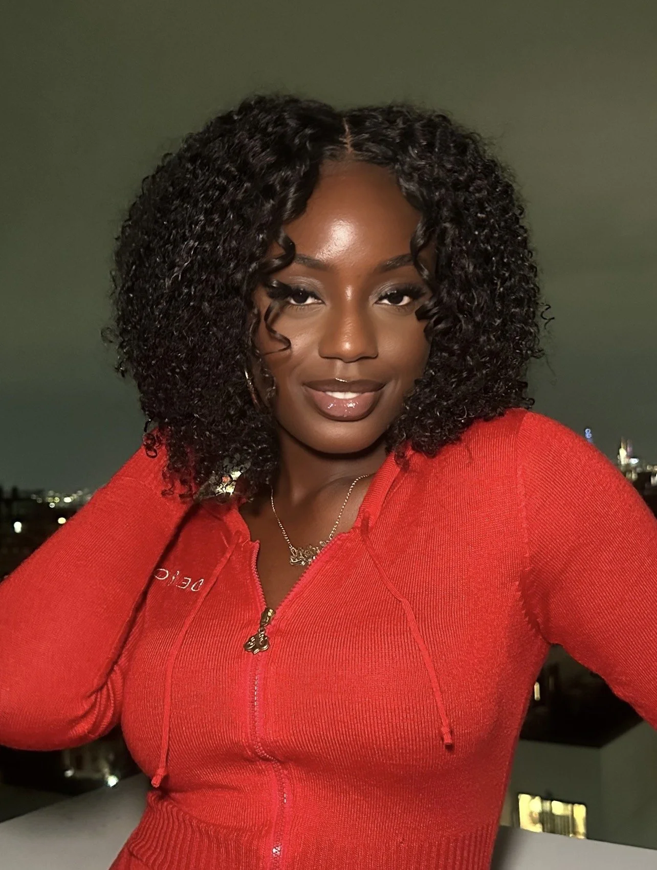 A woman with curly black hair wearing a red zip-up jacket, standing outdoors at night with a city skyline in the background.