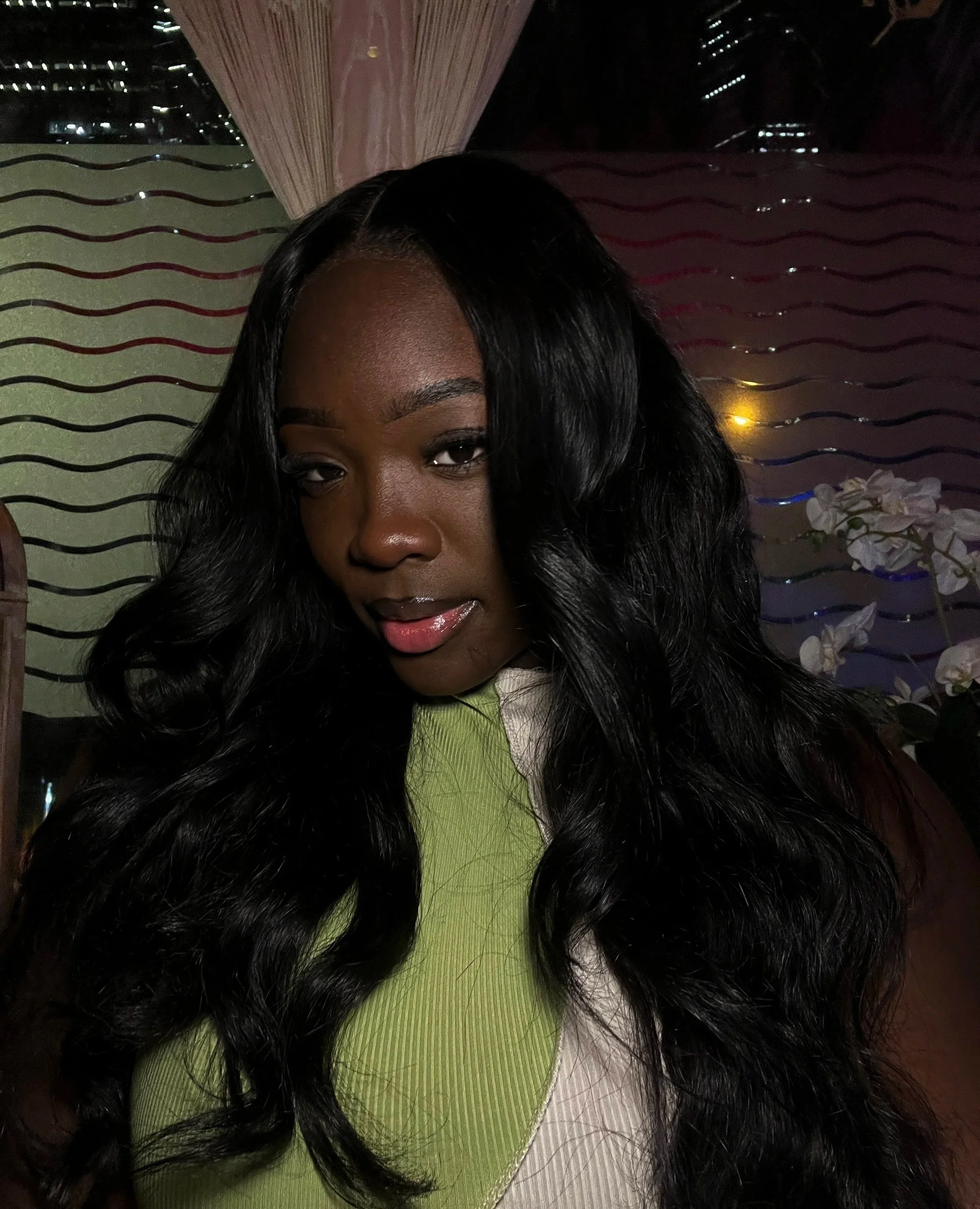 A woman with long black curly hair, wearing a lime green and beige top, posing indoors with a dark background and decorative wall featuring wavy stripes and floral accents.