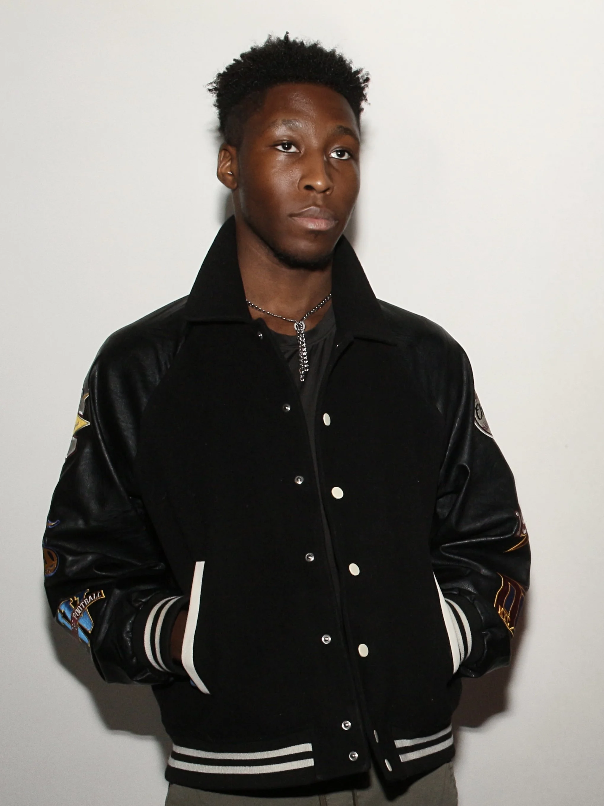 A young man with dark skin and short curly hair is standing against a plain white wall, wearing a black varsity jacket with patches on the sleeves and a silver chain necklace.