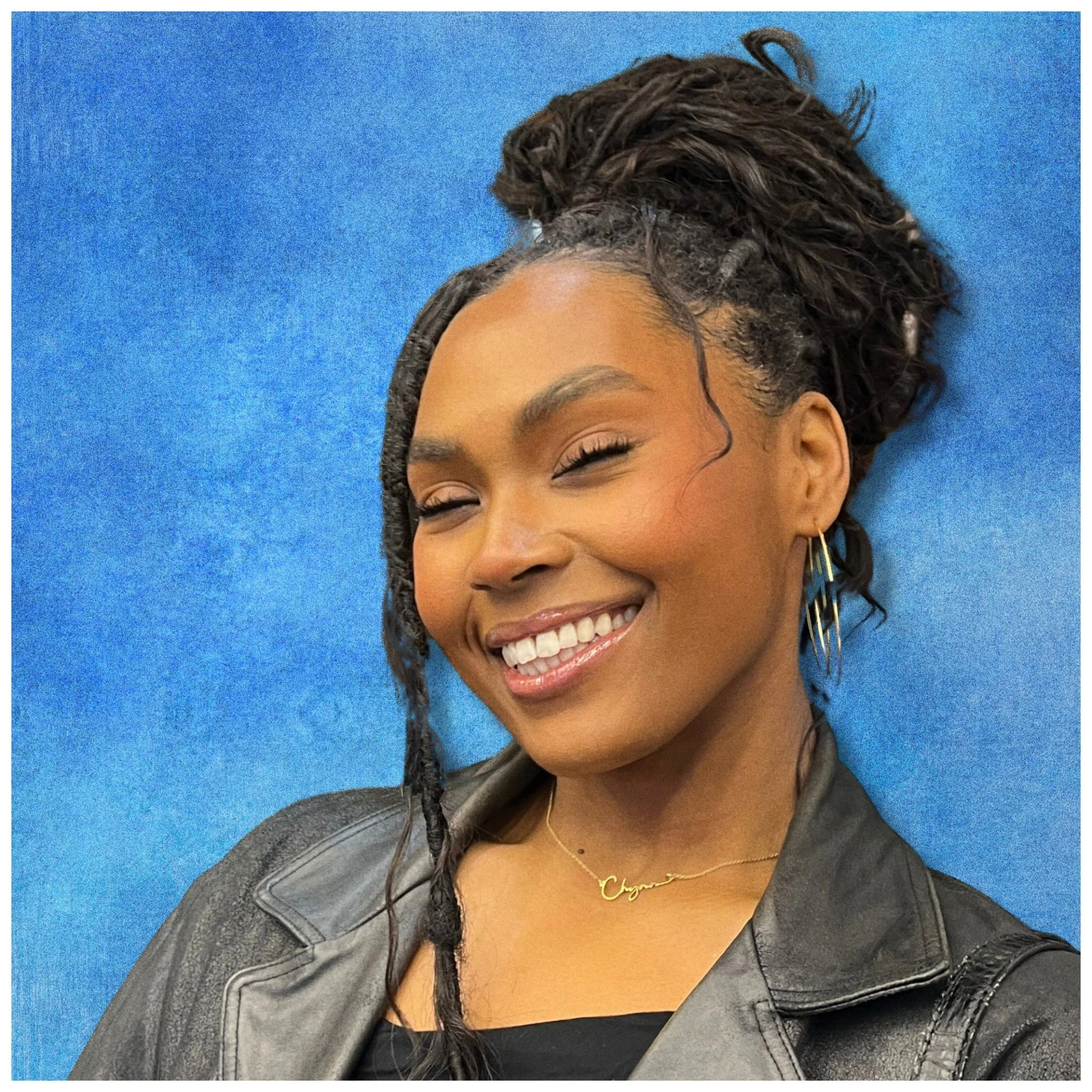 A smiling woman with dark skin, curly hair styled in dreadlocks, wearing earrings, a necklace, and a leather jacket, standing against a blue background.