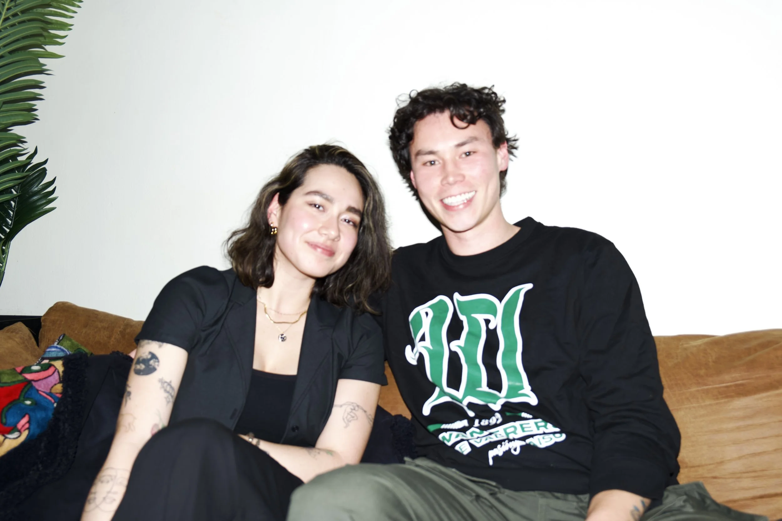 Two young people sitting on a brown sofa, smiling at the camera. The woman has dark curly hair, tattoos on her arms, and is wearing a black top and gold jewelry. The man has short, curly dark hair, and is wearing a black sweatshirt with green and white text and olive-green pants. There is a plant in the background and colorful pillows on the sofa.