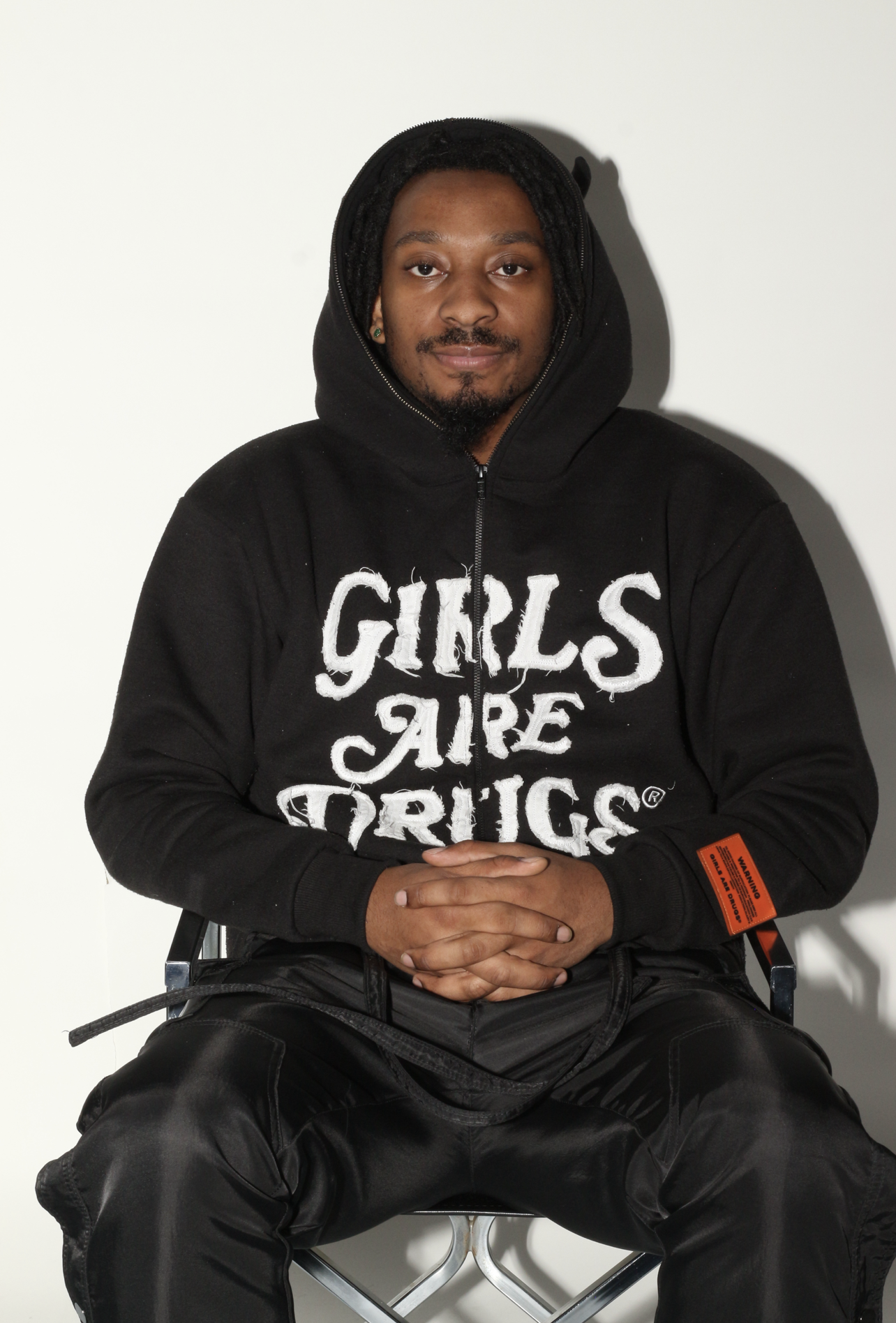 A man sitting on a chair, wearing a black hoodie with white text that reads 'GIRLS ARE DRUGS', and black pants. He has a beard, is smiling slightly, and has his hands clasped.