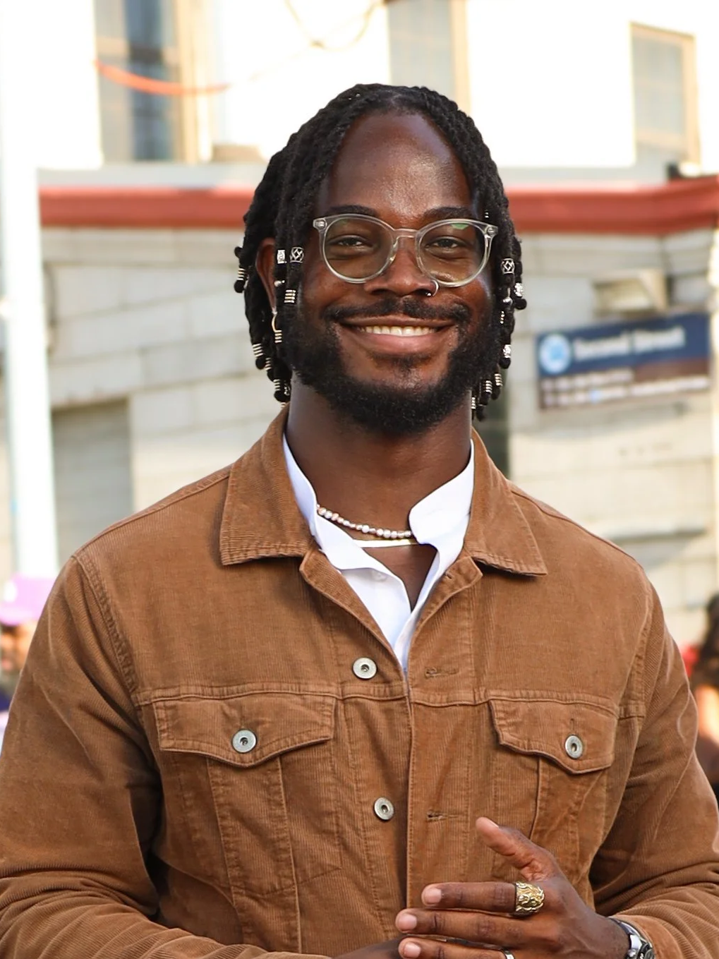 A joyful man with dreadlocks and glasses, smiling outdoors, wearing a brown jacket, white shirt, and accessories including a ring and necklace.