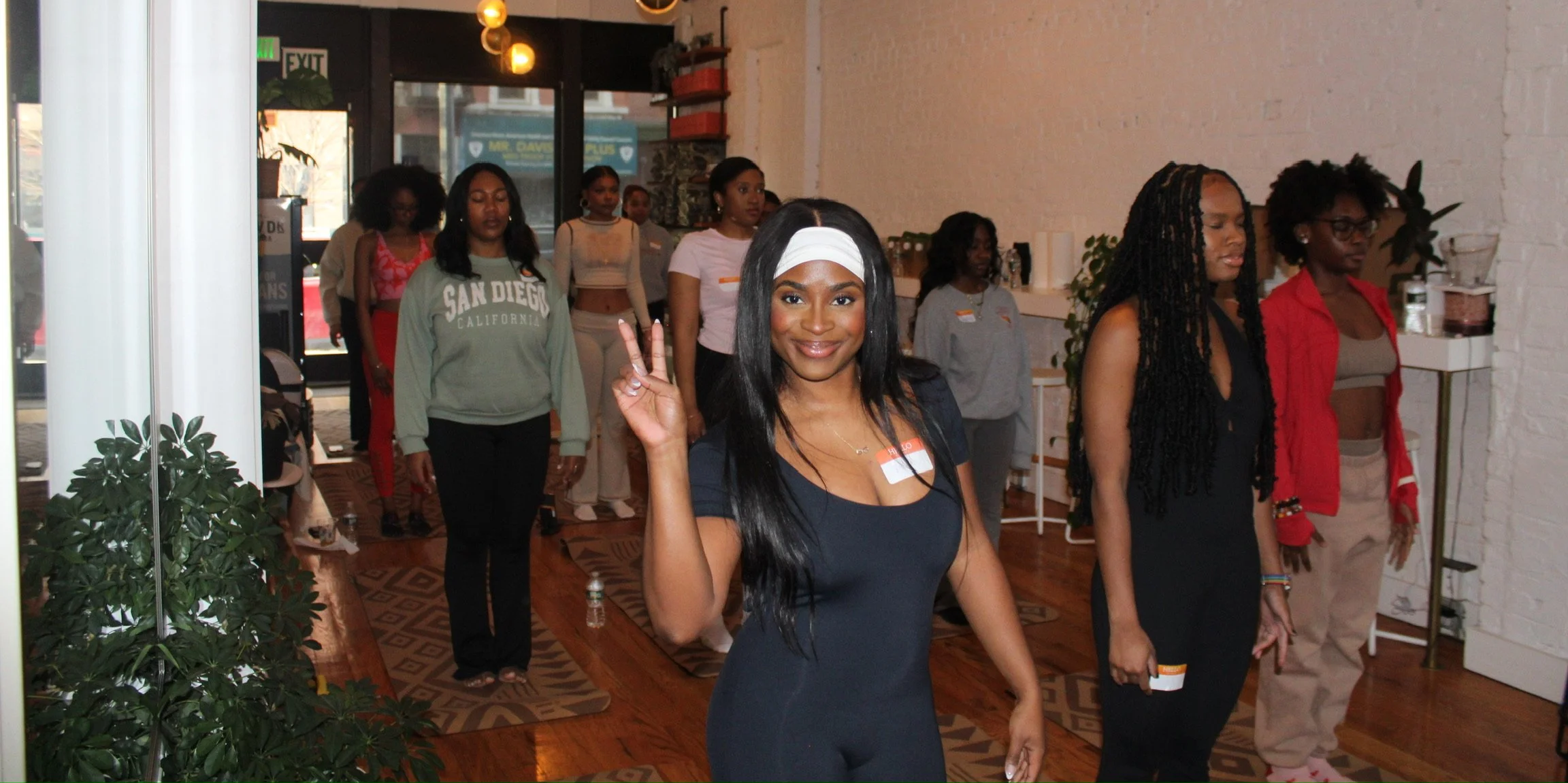 A group of women participating in a yoga or meditation class inside a studio, with one woman in the front making a peace sign and smiling at the camera.