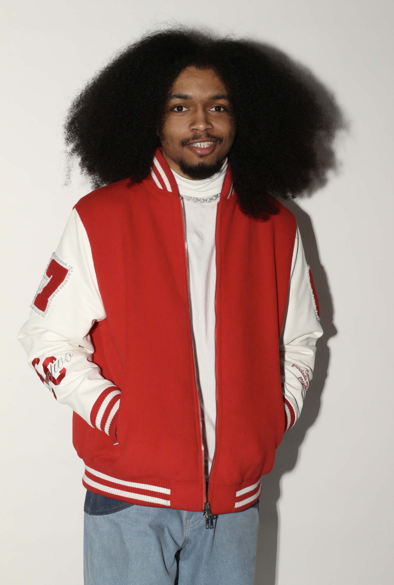 Young man with large, curly black hair wearing a red and white varsity jacket and light blue jeans, standing against a plain white background.