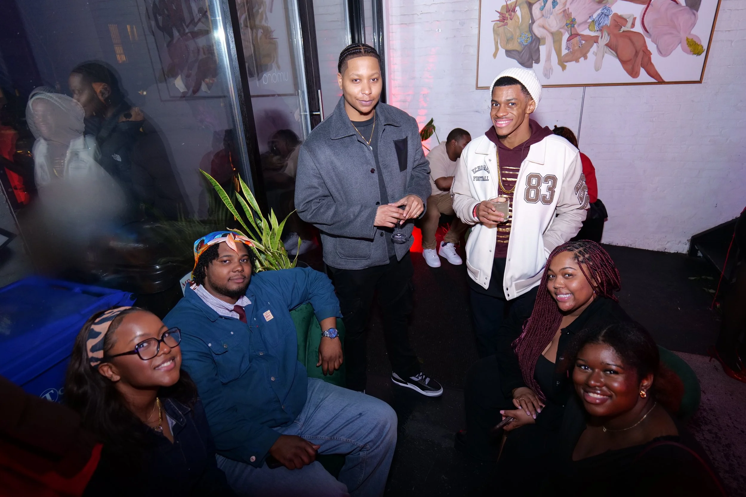 A group of six young adults at a social gathering inside a venue with white brick walls and modern art. They are smiling and holding drinks, with some seated and some standing. The setting appears casual and lively.