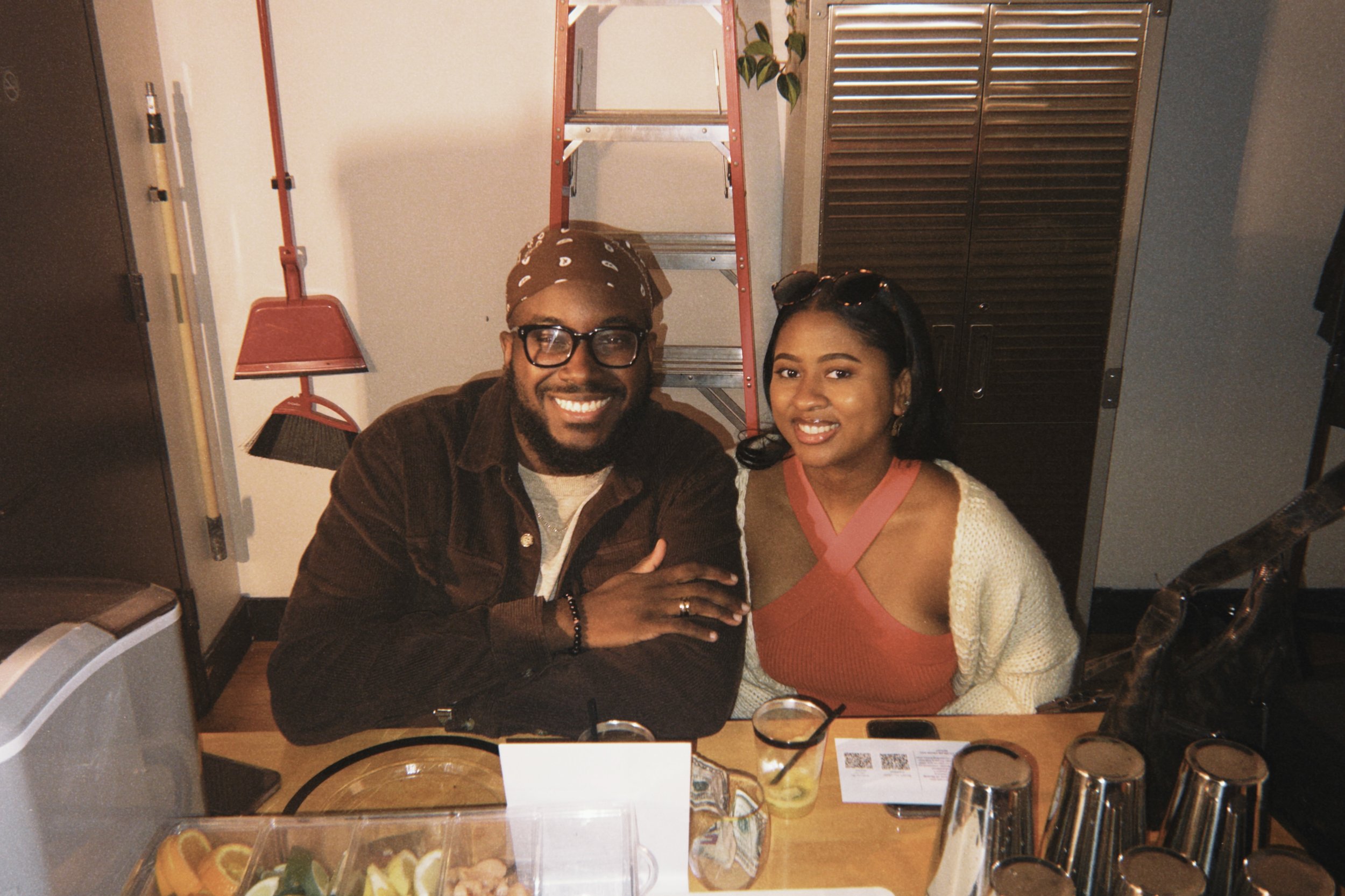 A smiling man and woman sitting at a counter in a casual setting, with various bar tools and drinks in front of them.
