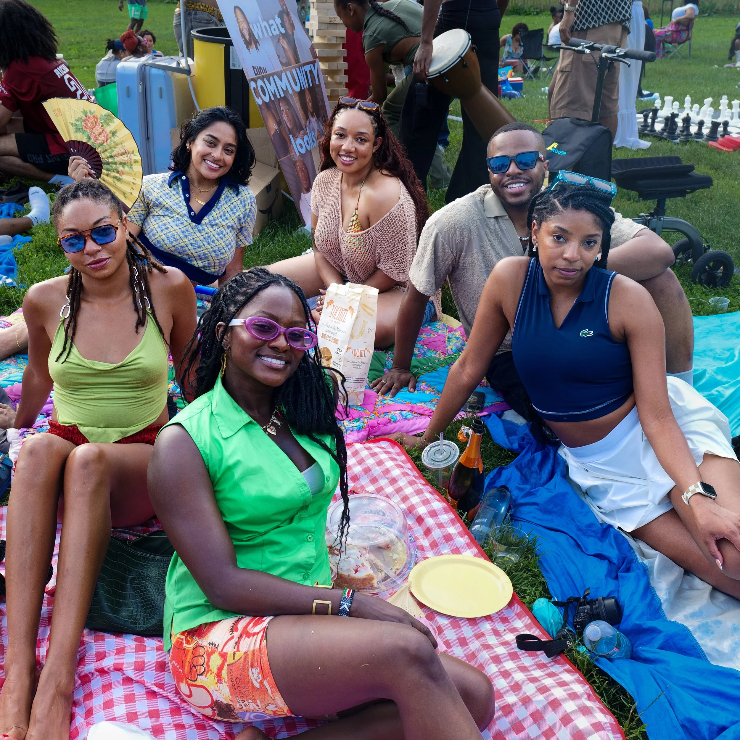 A group of friends enjoying a picnic outdoors on a sunny day, sitting on a red and white checkered blanket with food and drinks, surrounded by other people, some playing chess and others relaxing on the grass.