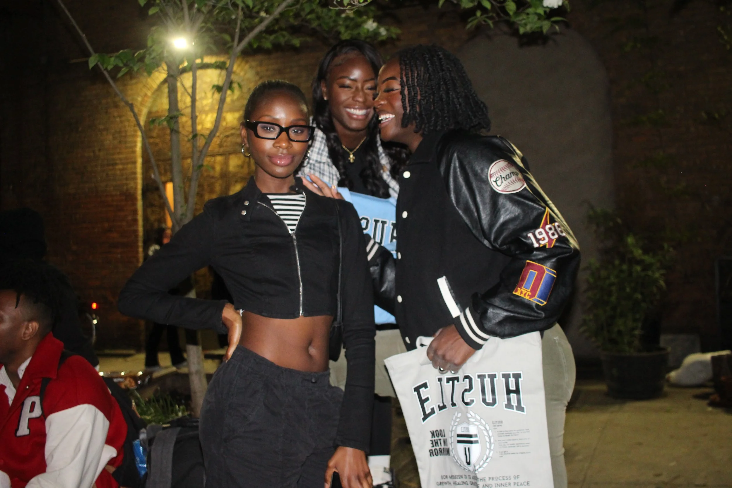 Three women standing together at night outdoors, smiling and posing for the camera. One woman wears glasses and a black zip-up jacket with a striped shirt underneath, another in the middle has a big smile and wears a hoodie with a jacket, and the third woman on the right has curly hair and is wearing a black jacket with patches, holding a white bag.