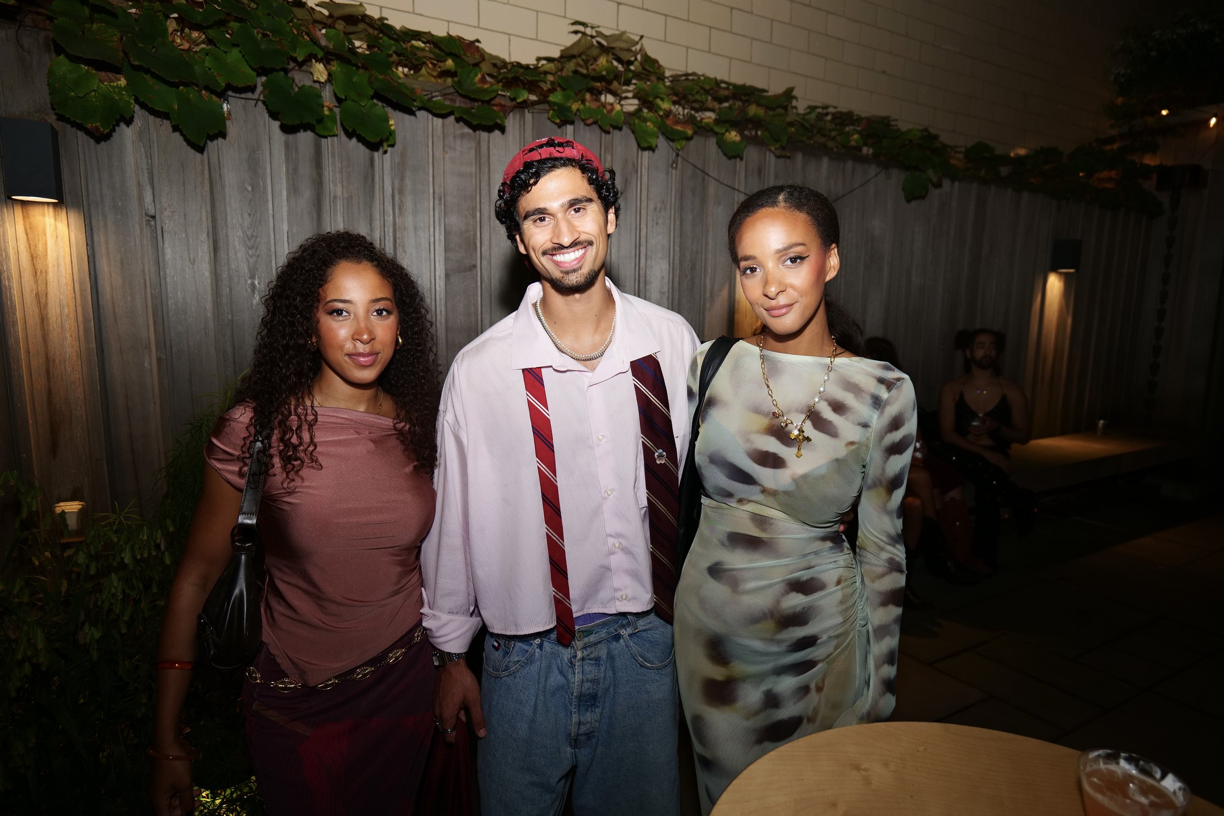Three people at a social gathering, two women and one man, standing in front of a wooden fence with greenery. They are smiling and dressed stylishly.