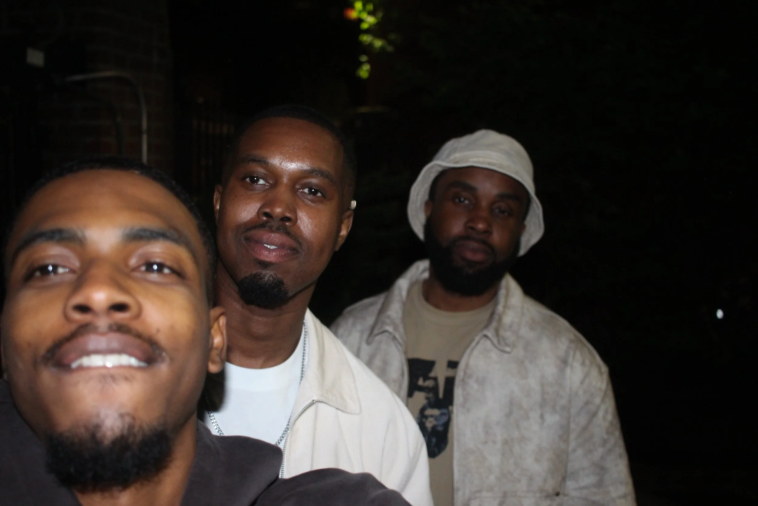 Three men posing for a selfie outdoors at night, with a dark background and some greenery visible.