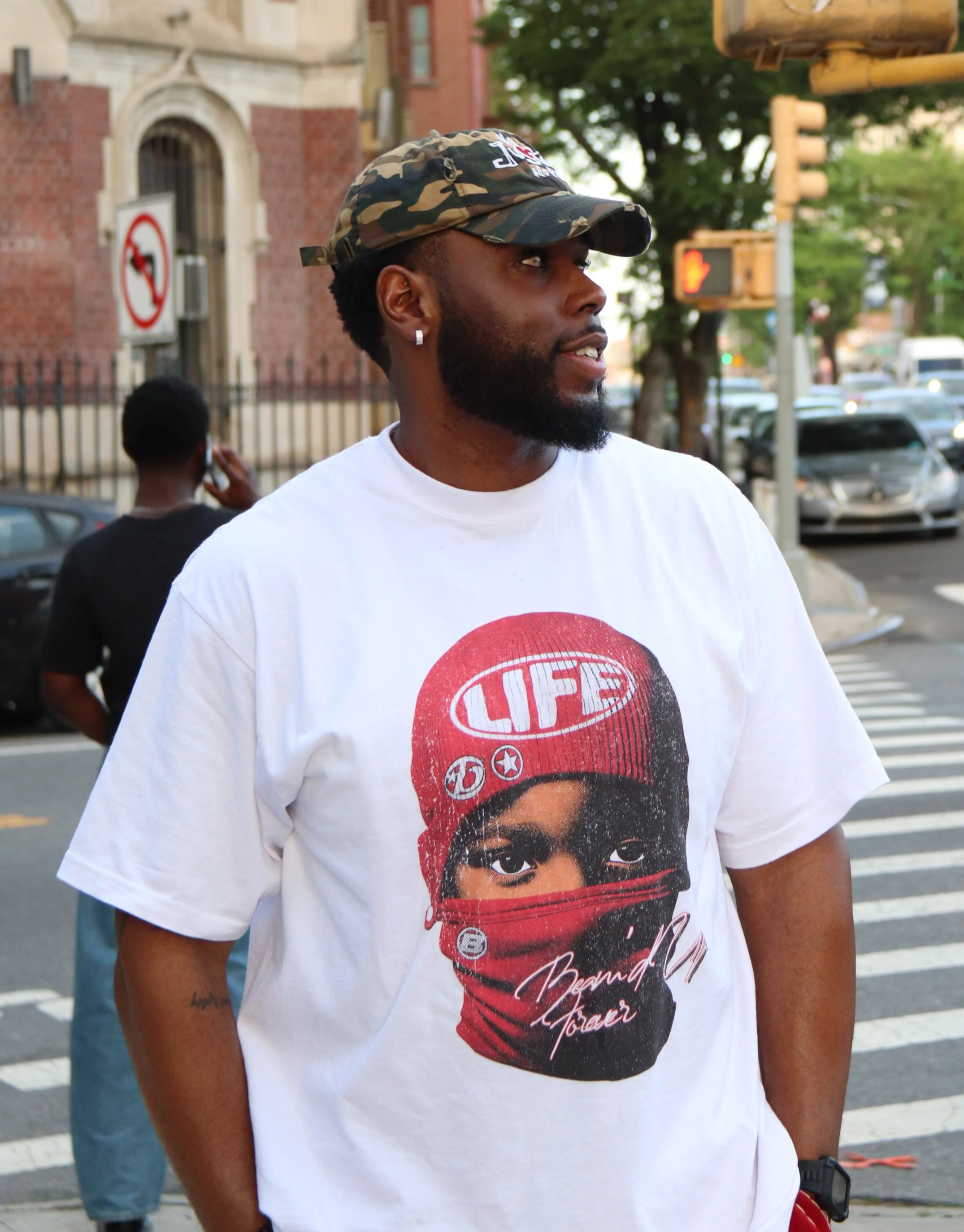 A man with a beard and earrings wearing a camouflage cap and a white t-shirt standing at a crosswalk on a city street, with cars and a person on the phone in the background.