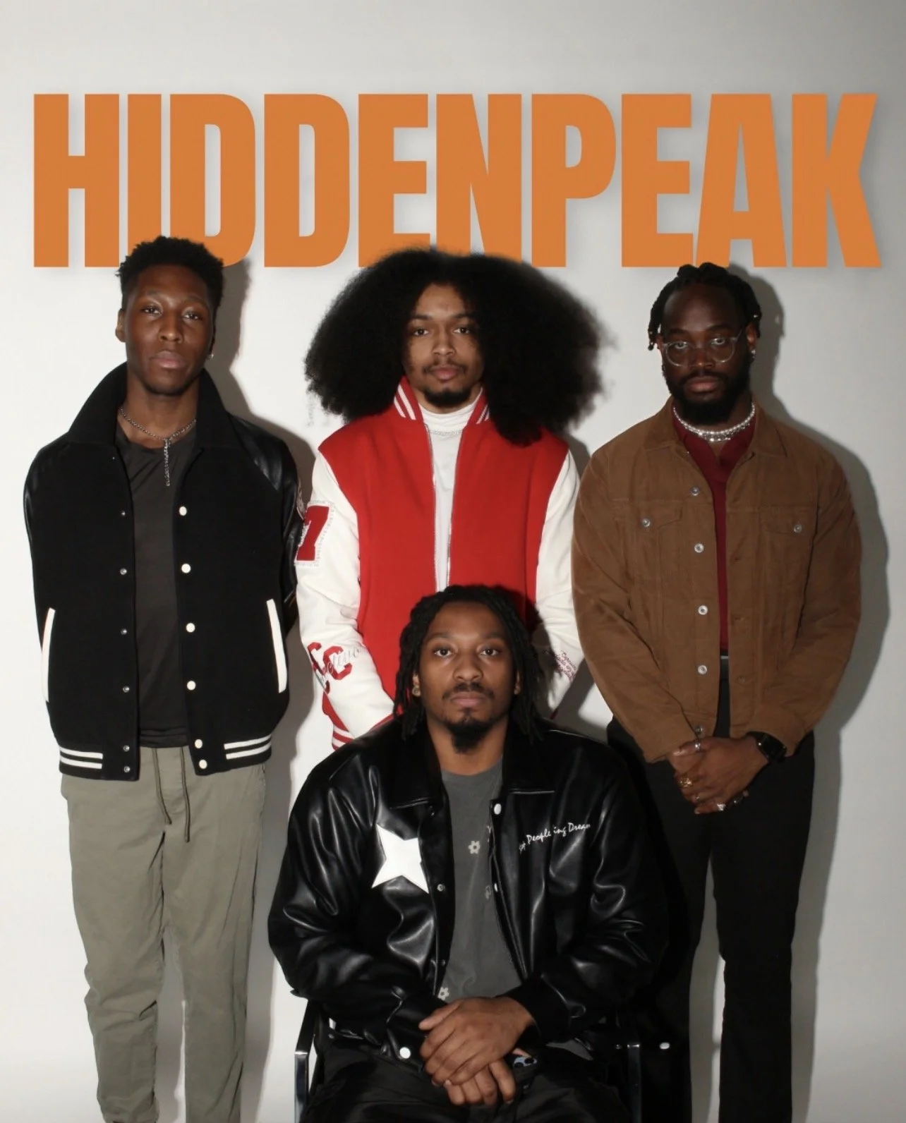 Five men posing for a photo in front of a wall with the word 'HIDDENPEAK' in large orange letters. One man is seated in front, and four men are standing behind him.