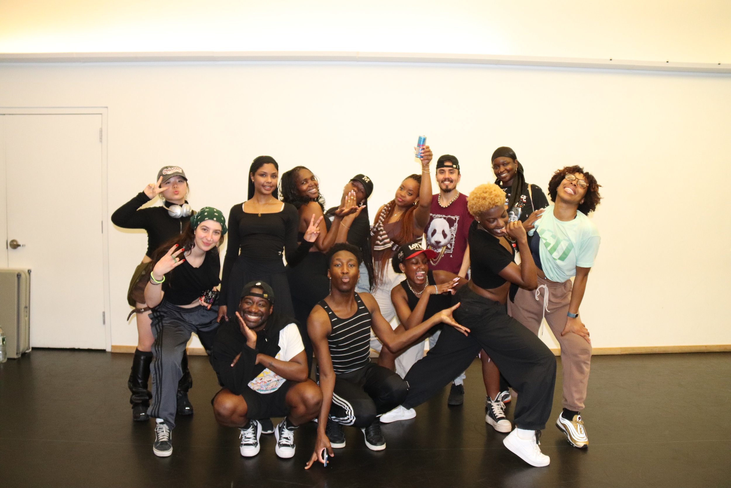Group of diverse young adults posing together in a dance studio or event space, some making peace signs and smiling, others in playful poses.